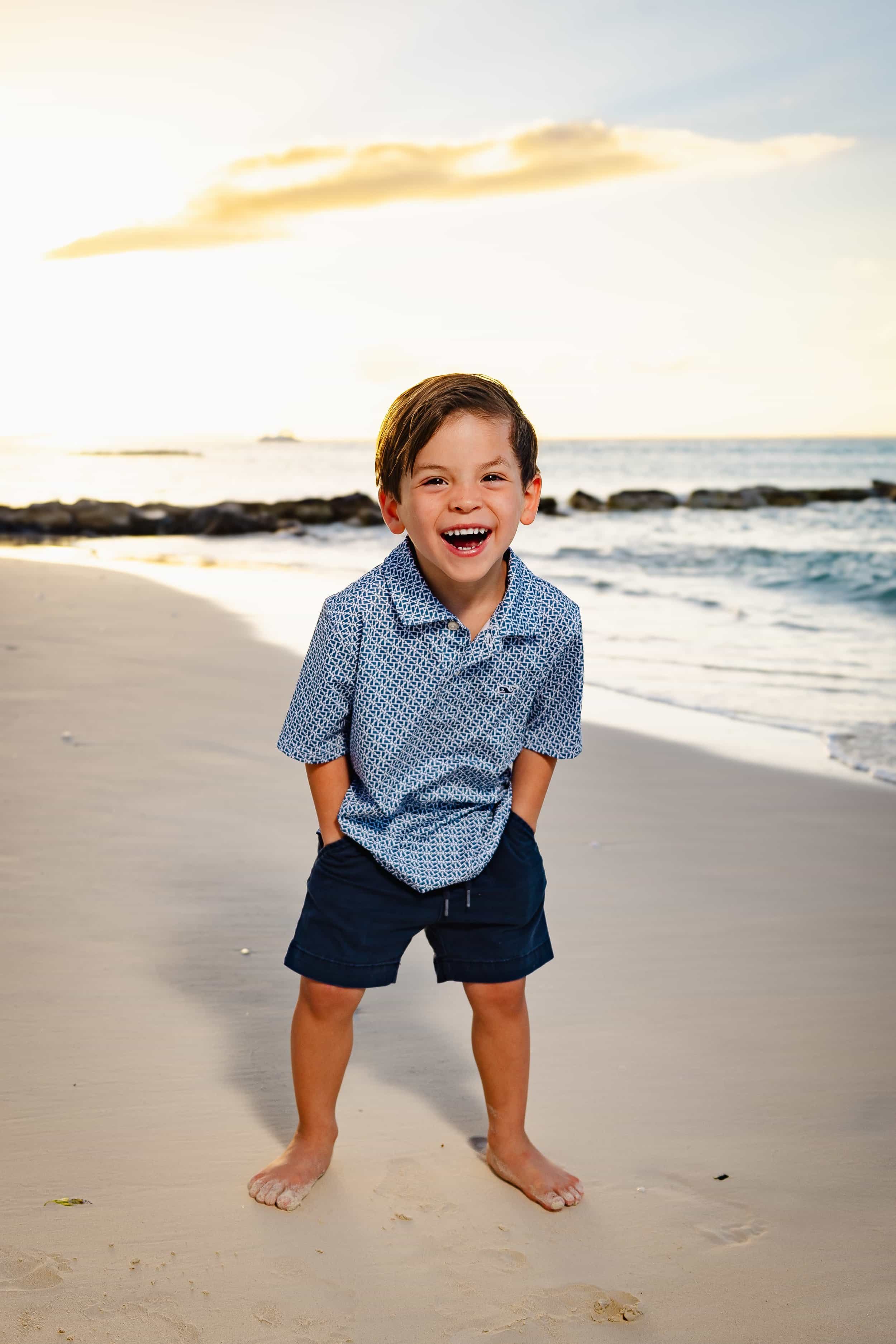 A young boy smiling and laughing on the beach at sunset, wearing a blue patterned short-sleeve shirt and navy shorts, with his hands in his pockets.