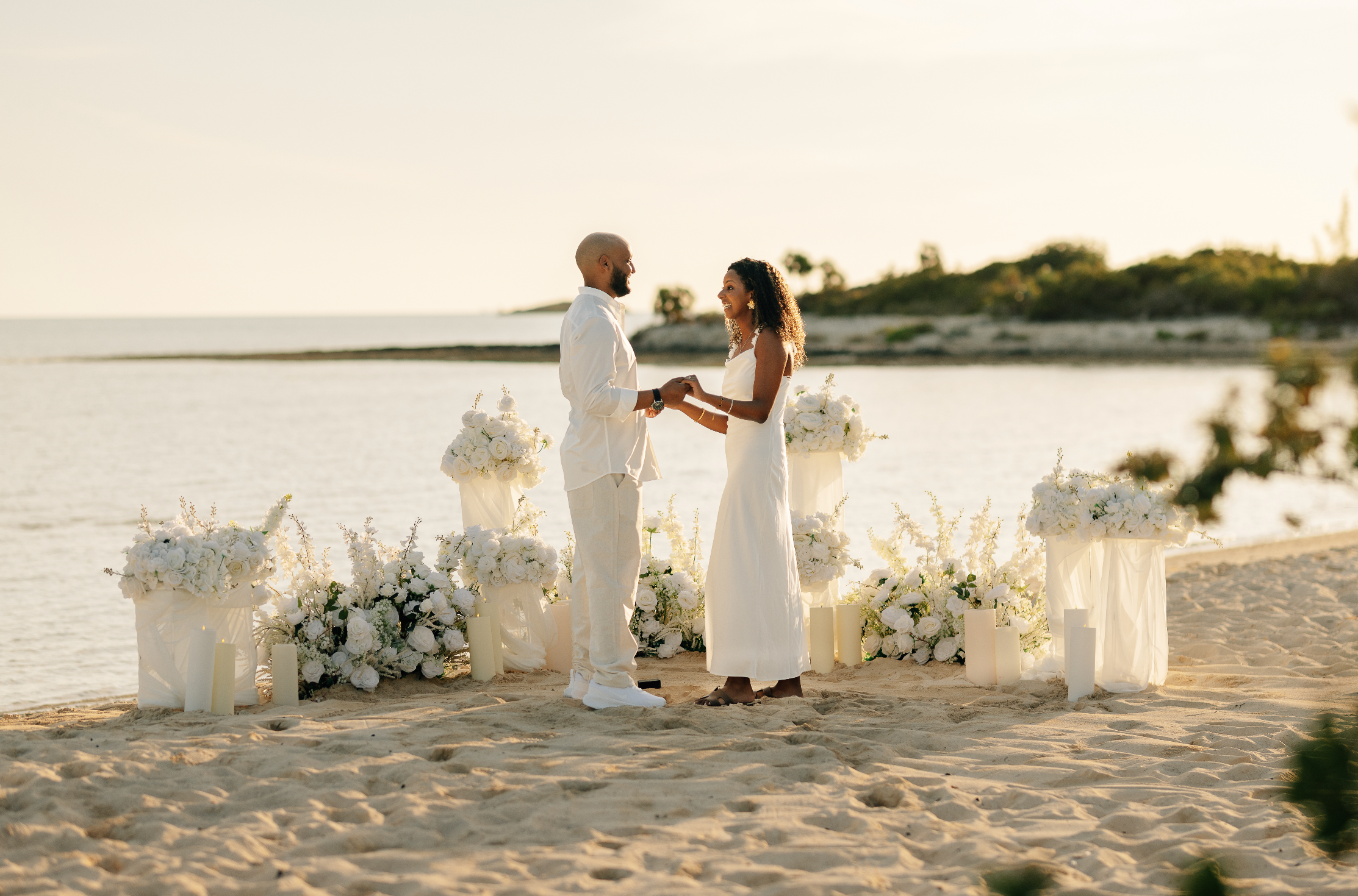 A couple exchanging wedding vows on a beach during sunset, with white floral arrangements and candles surrounding them.