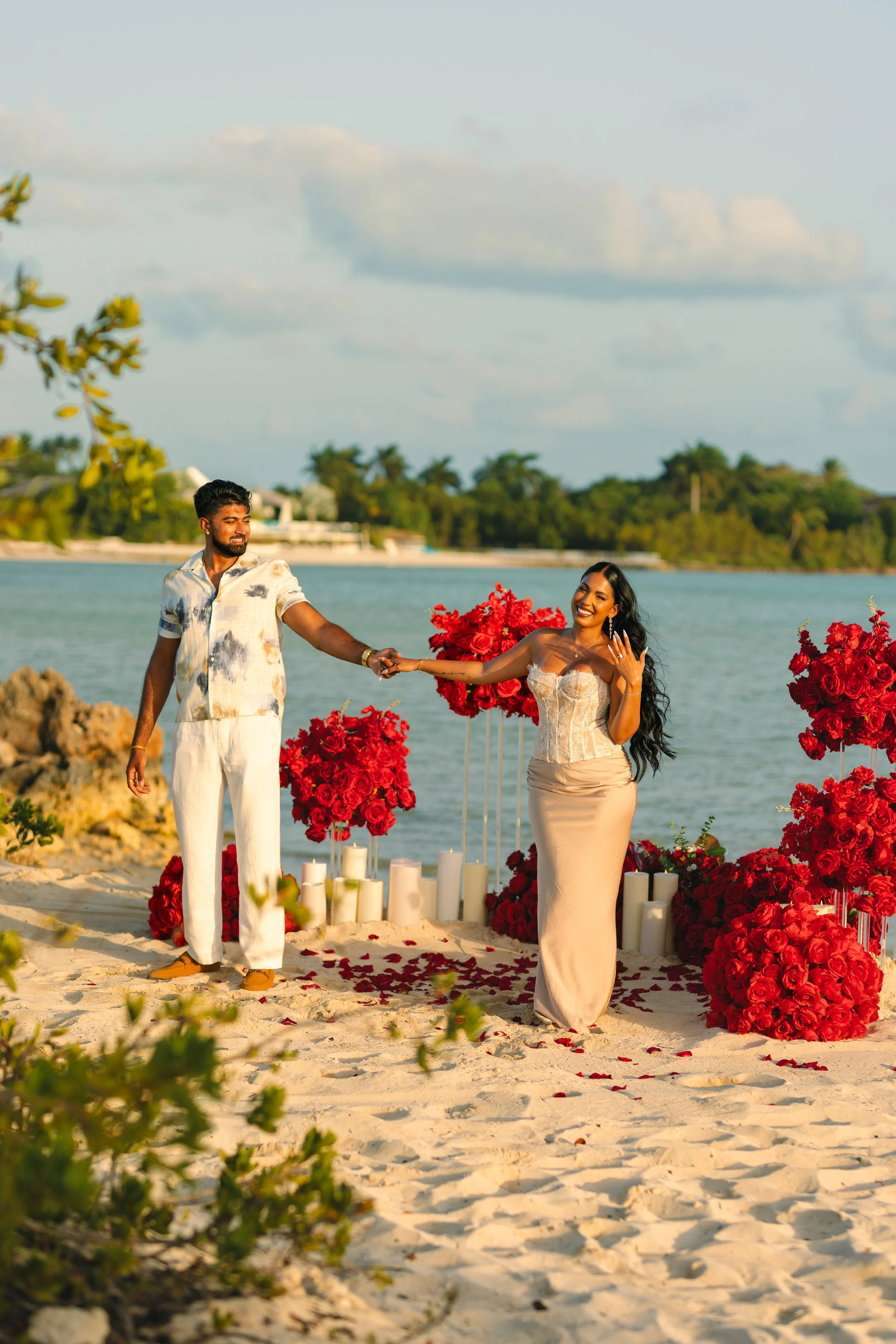 A couple holding hands on a sandy beach, surrounded by red roses, candles, and a decorative arch, with water and a forested shoreline in the background, during sunset.
