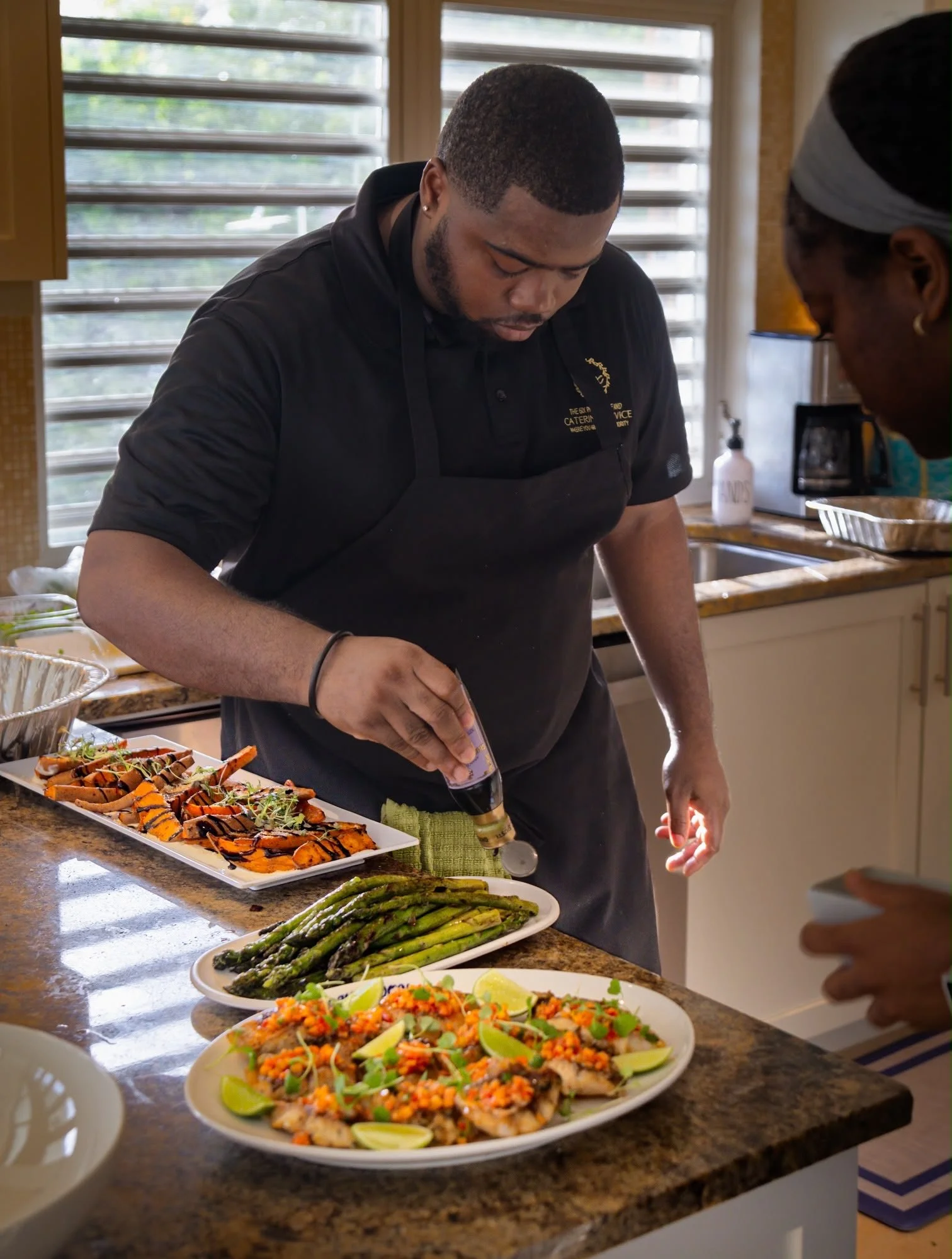 A man is preparing food in a kitchen with an assortment of dishes including grilled asparagus, a colorful salad with lime wedges, and a platter of grilled salmon topped with herbs and garnishes. There is another person partially visible on the right 