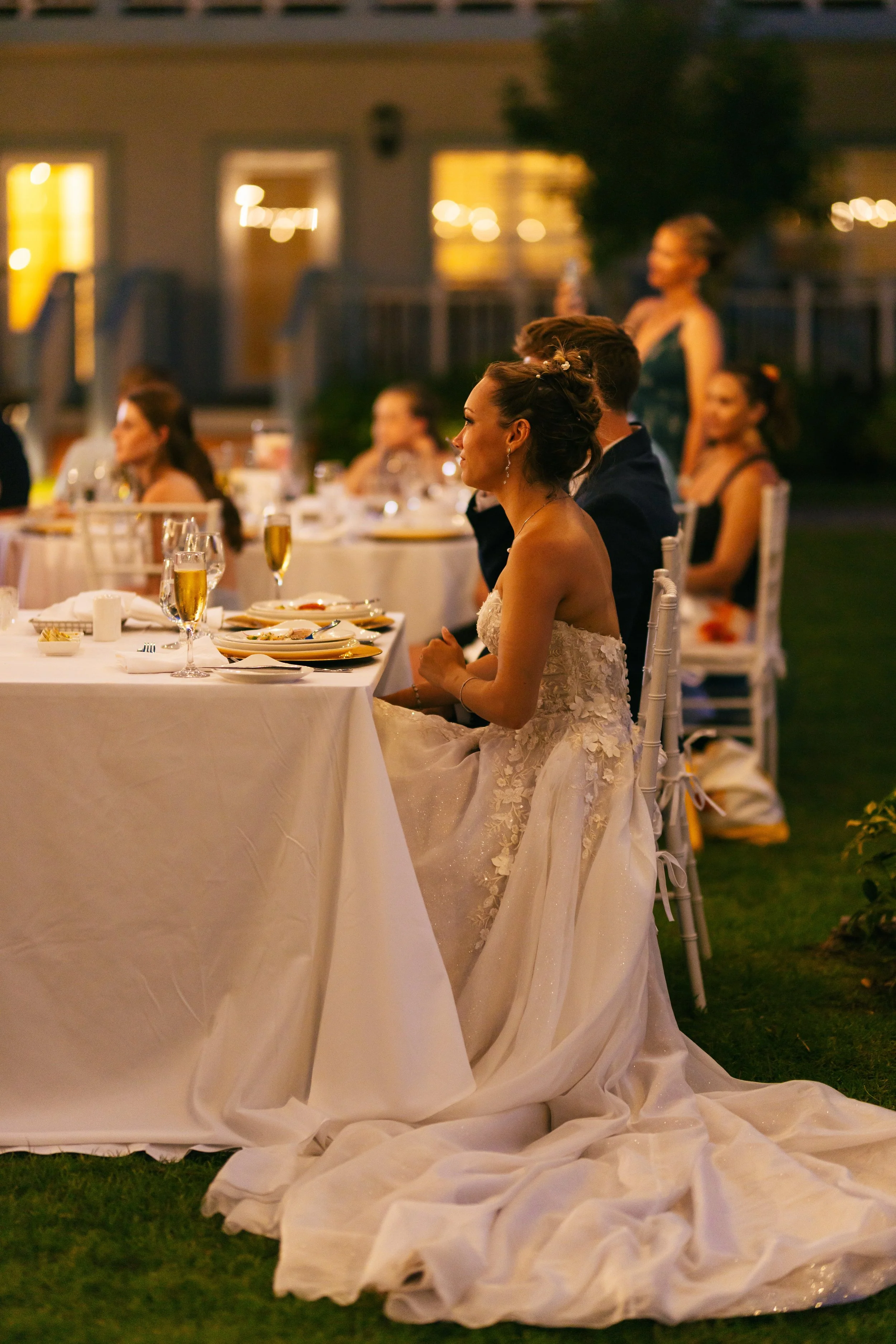 A bride sitting at a dinner table during a wedding reception, with a long train of her wedding dress flowing on the grass beneath. She is wearing a strapless lace wedding gown, with her hair styled up and wearing earrings. The setting appears to be o