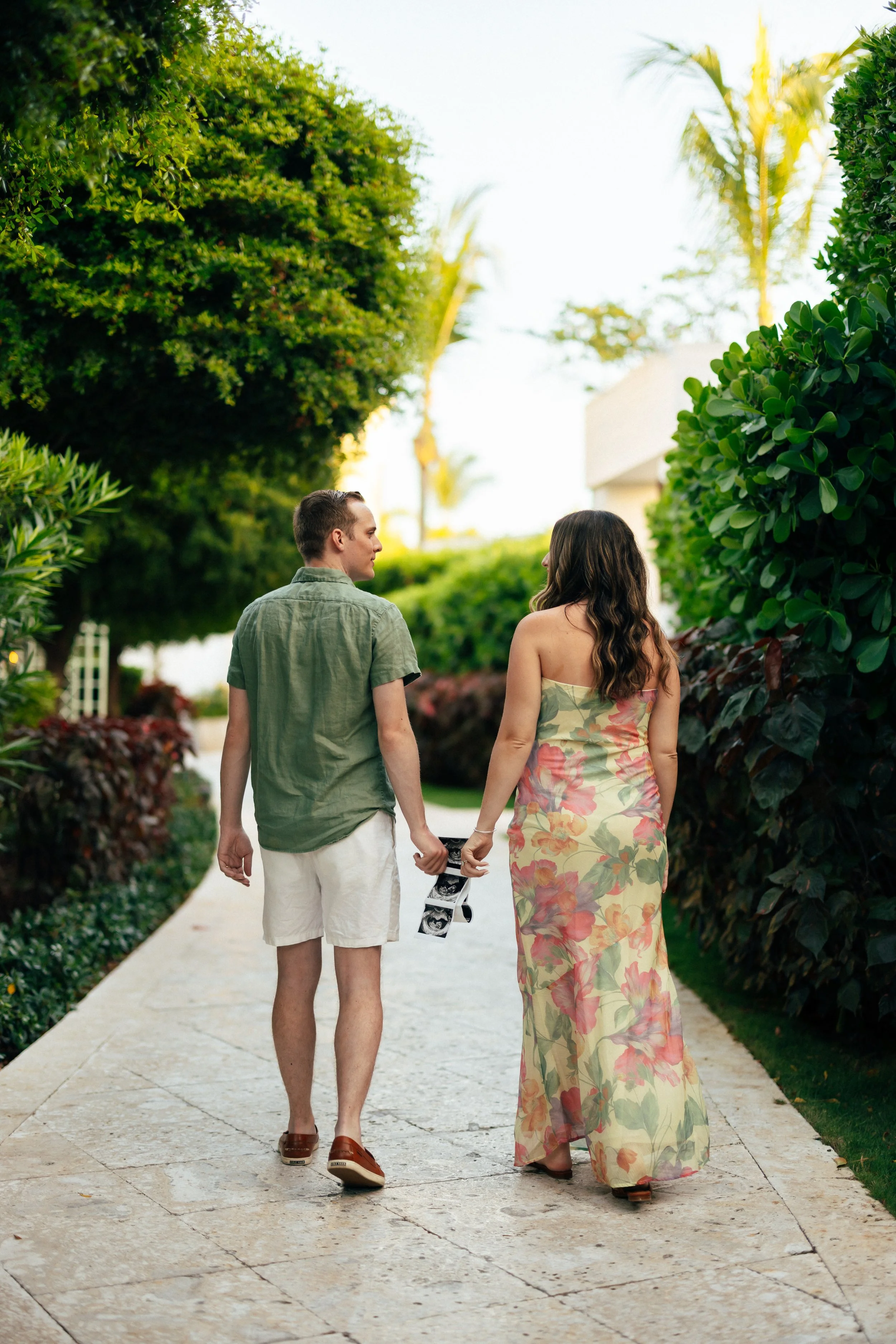 A couple walking hand in hand outdoors on a paved path surrounded by lush green bushes and trees, during daytime.