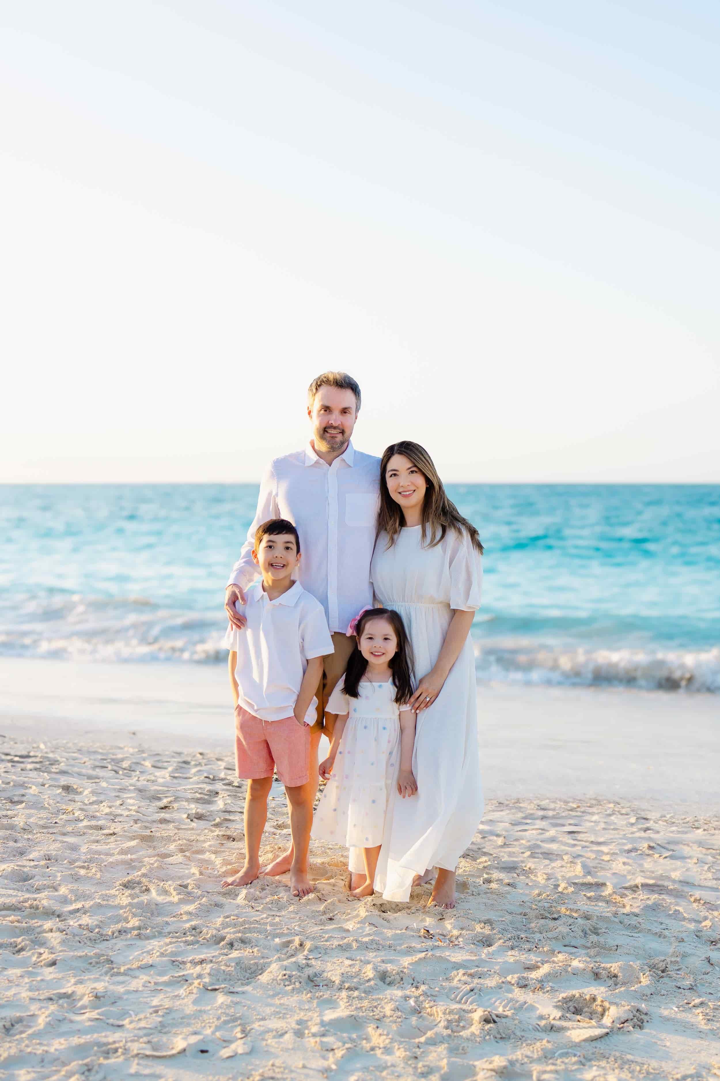 A family of five standing on the sandy beach near the ocean, dressed in white and light-colored clothing, smiling for a photo with the ocean and sky in the background.