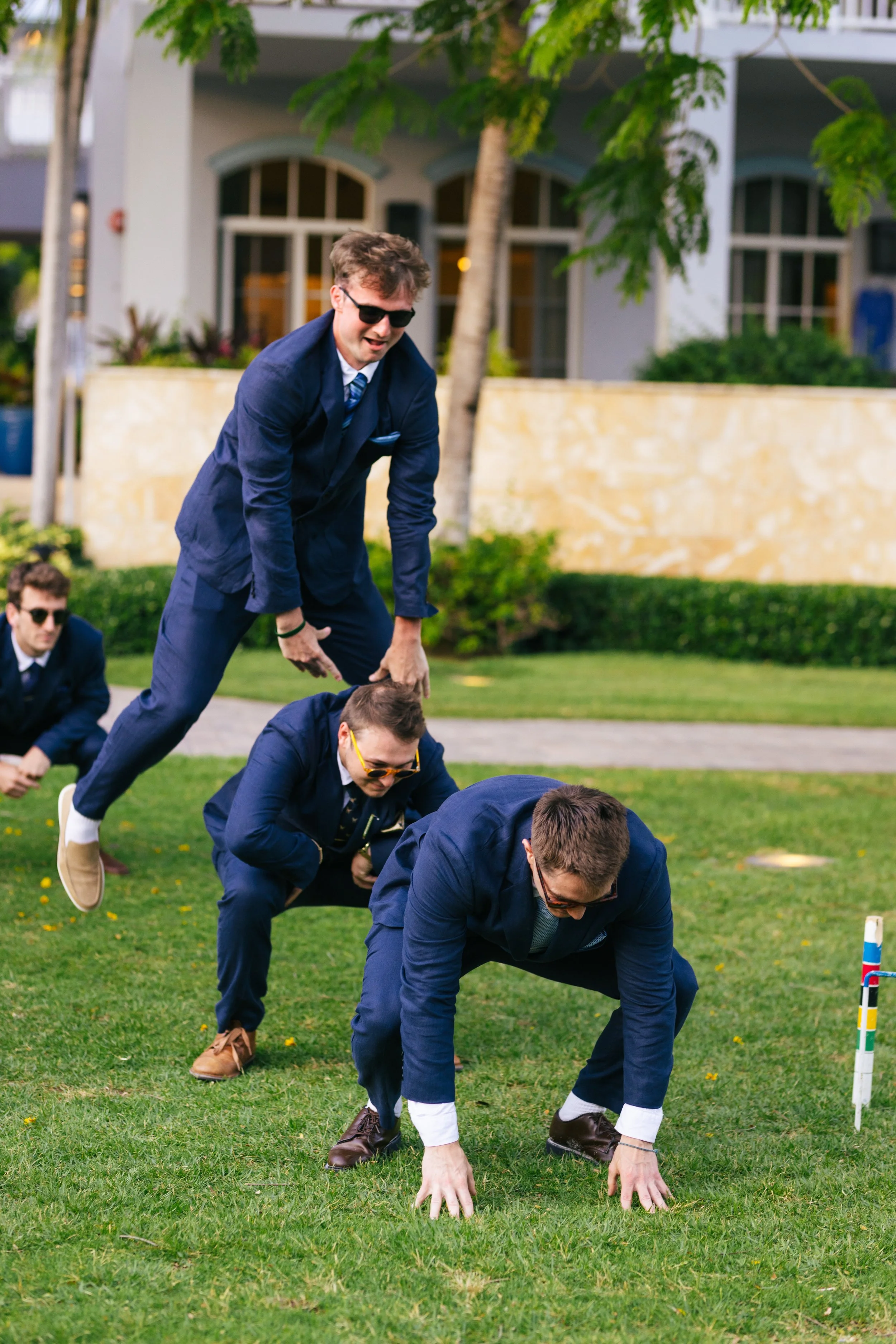 A group of young men wearing suits and sunglasses are participating in a conga line, crawling on their hands and knees on a grassy lawn in an outdoor setting with trees and a building in the background.