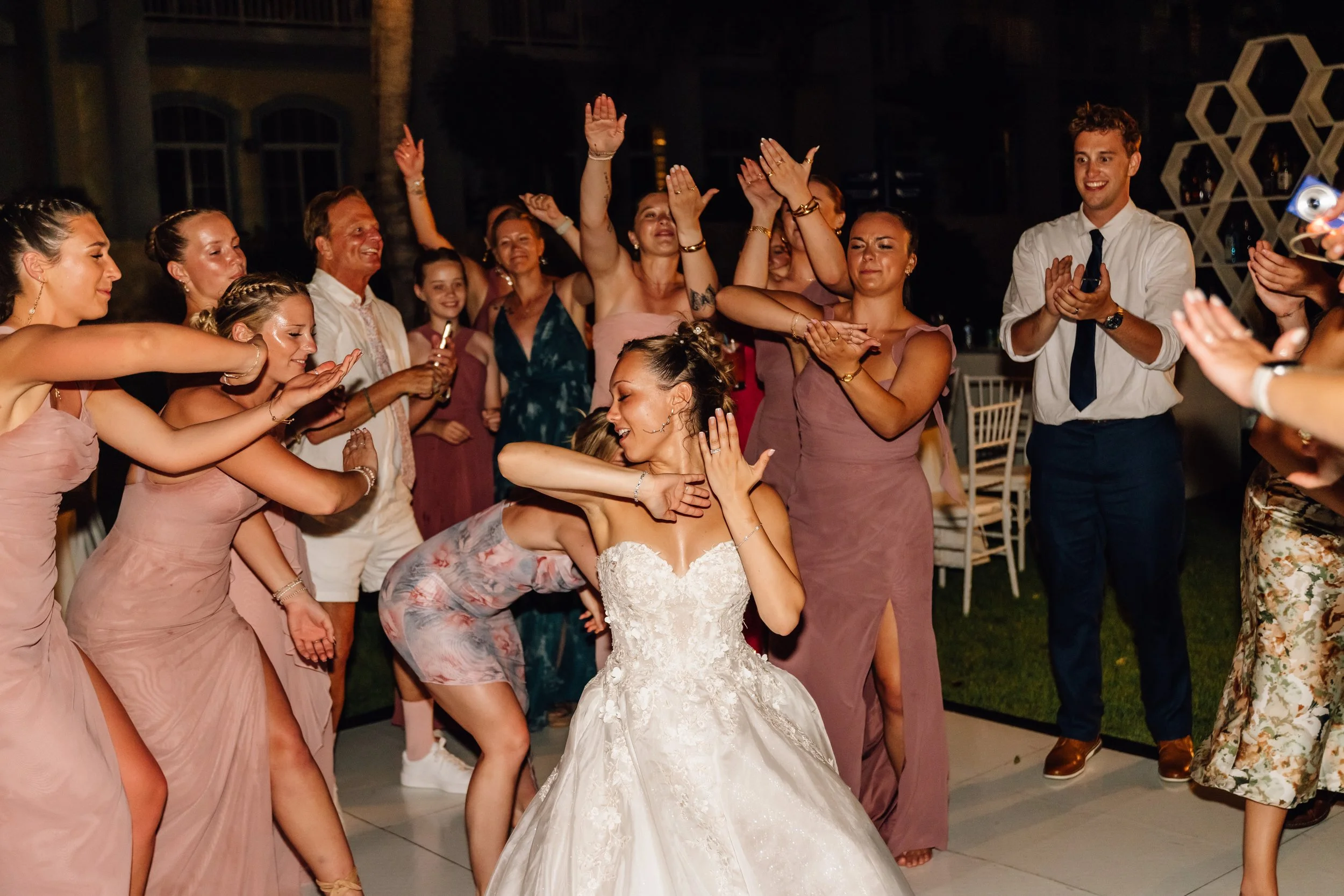 A wedding celebration with a bride in a white wedding gown dancing among a group of women in pink dresses and other guests, all smiling and enjoying the moment.