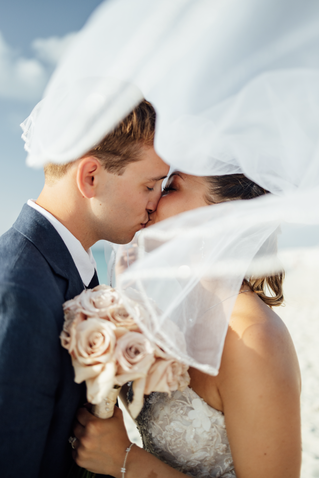 A bride and groom sharing a kiss, with the bride holding a bouquet of pale pink roses, under the bride's veil on a beach with cloudy sky.