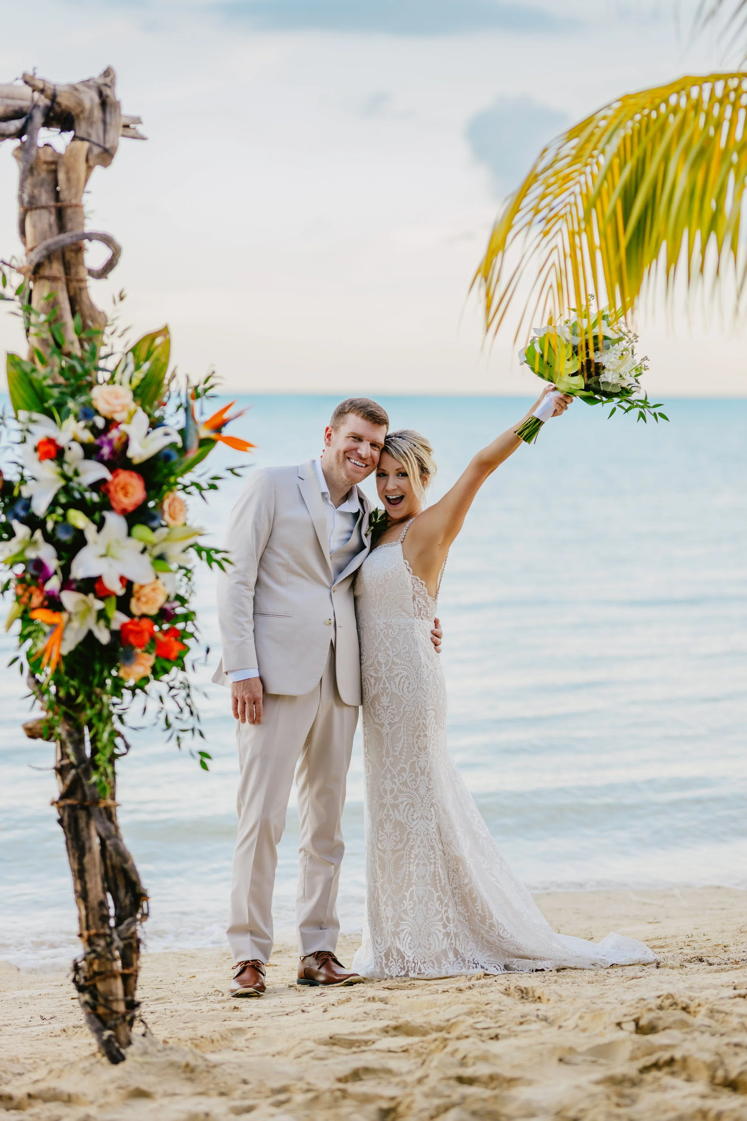 A happy bride and groom in wedding attire standing on a sandy beach with the ocean in the background. The bride is holding a bouquet of flowers and raising her arm, while the groom is smiling and standing close to her.