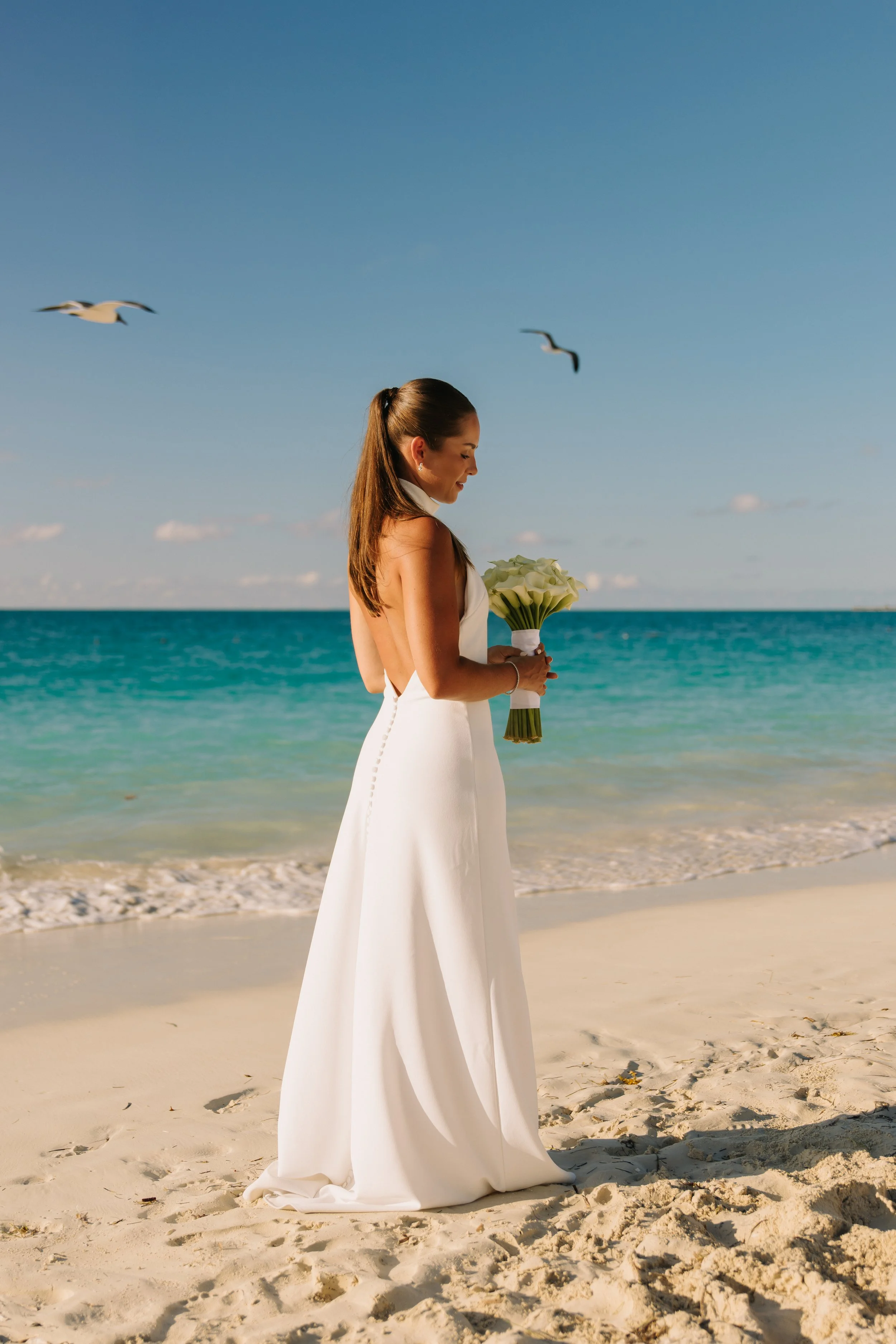 A woman in a white dress standing on the beach, holding a bouquet of white calla lilies, with the ocean and blue sky in the background, and two seagulls flying overhead.