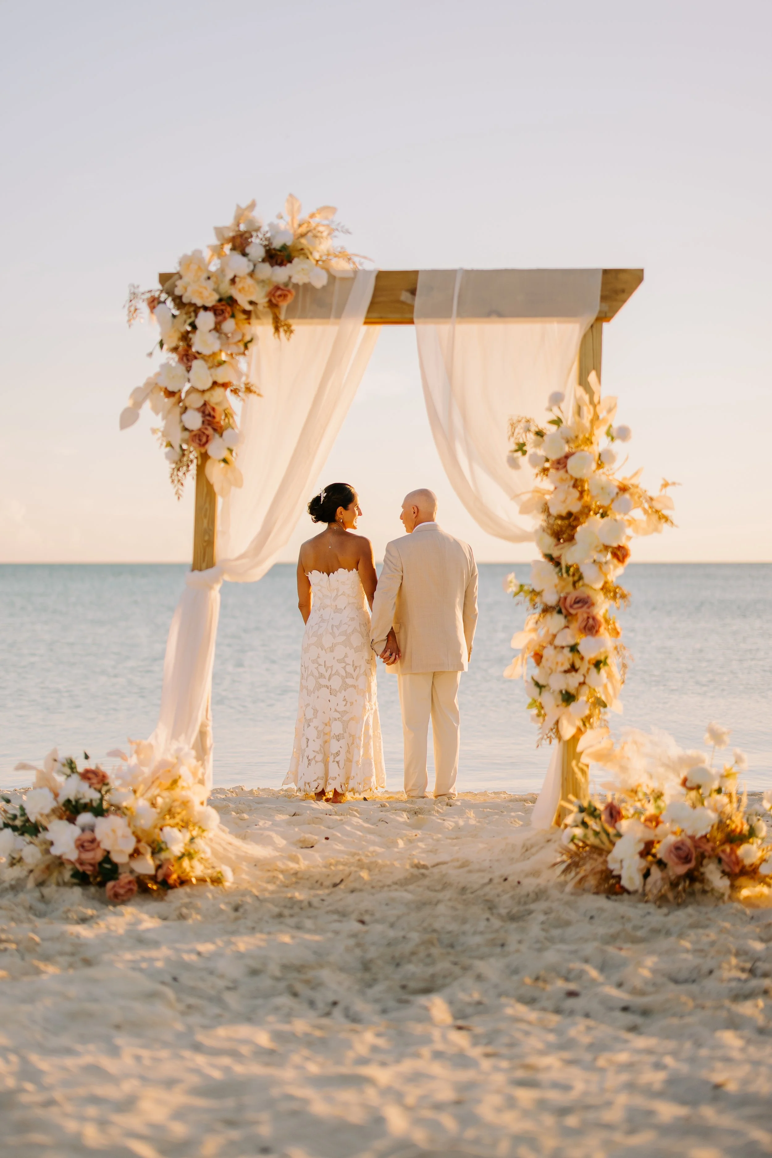 A wedding ceremony is taking place on the beach with a wooden arch decorated with white and blush-colored flowers and sheer white drapery. The bride and groom are holding hands, standing in front of the arch, with the ocean and a clear sky in the bac