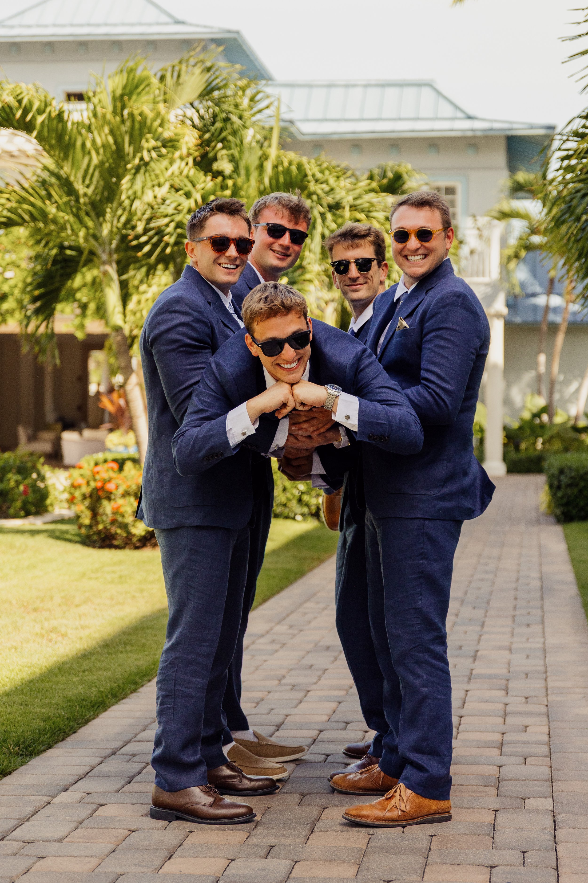 A group of six men in blue suits and sunglasses, smiling and posing together outside on a paved walkway with greenery and a large house in the background.