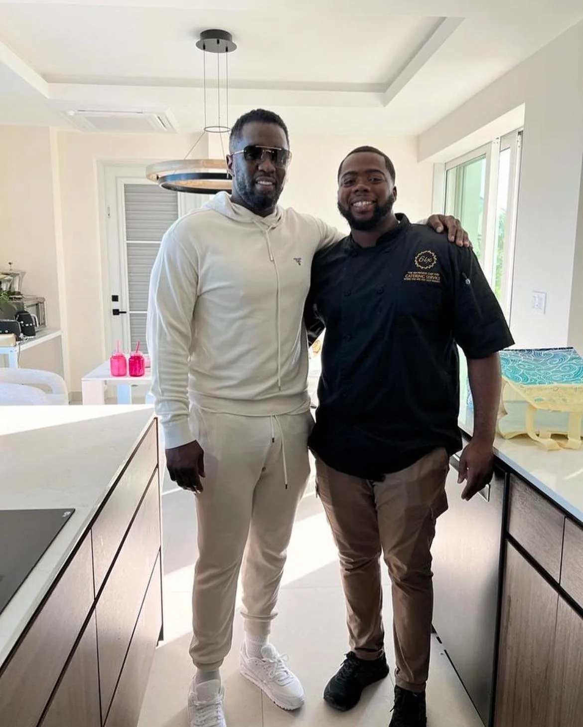 Two men standing in a modern kitchen, smiling with their arms around each other. One is wearing a white hoodie and pants with sunglasses, the other is wearing a black chef's coat and beige trousers.