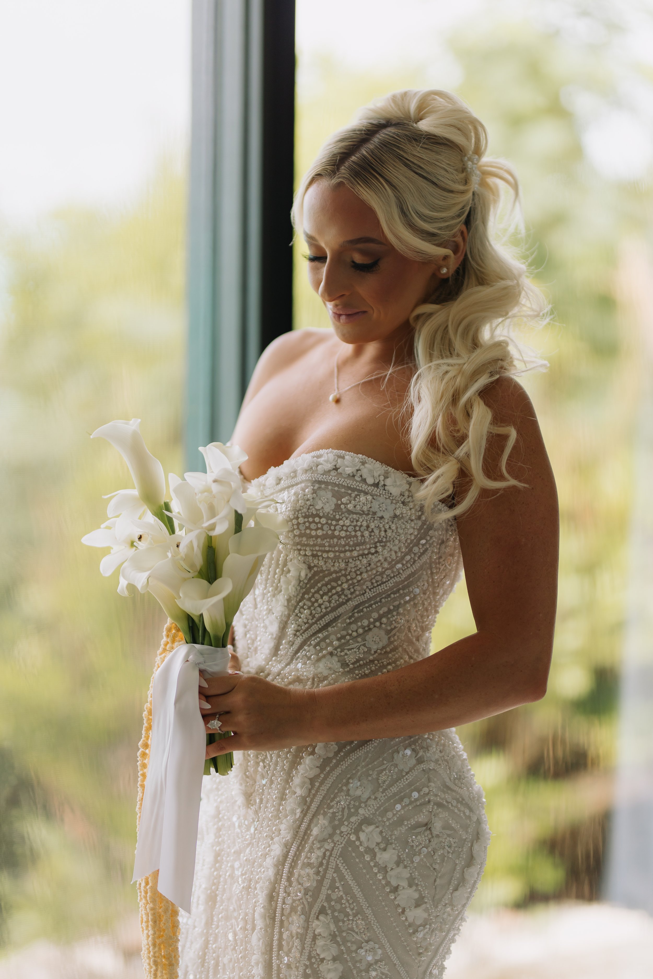 A bride with platinum blonde hair styled in loose curls, wearing a white wedding gown with intricate beading, holding a bouquet of white calla lilies, standing near a window with greenery outside blurred in the background.