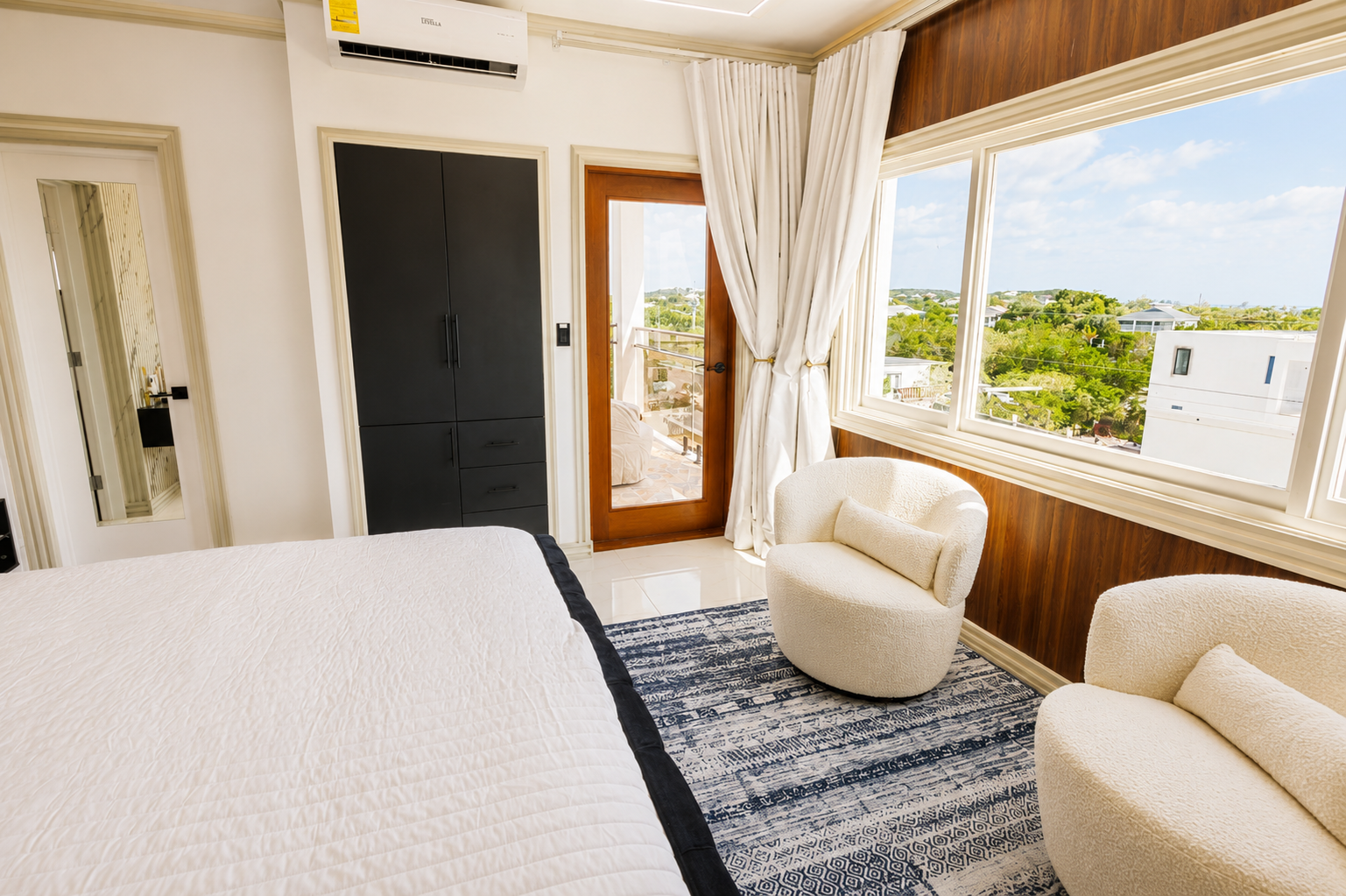 A bright bedroom with large windows showing a view of trees and houses outside, a white bed, a black wardrobe, a small balcony, and two cream-colored armchairs with pillows.