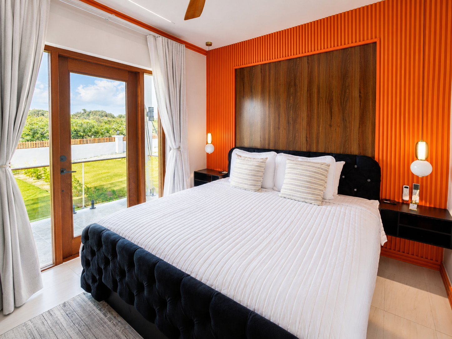 A bedroom with a large bed, decorative pillows, and a wooden accent wall. There are glass doors opening to a balcony with outdoor greenery visible through the windows, and white curtains hang on either side of the doors. Two modern wall-mounted lamps