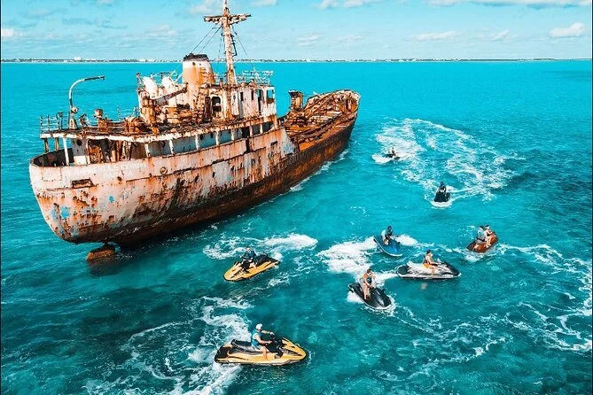 Rusty shipwreck near the coast with several jet skis and a small boat nearby on turquoise water.