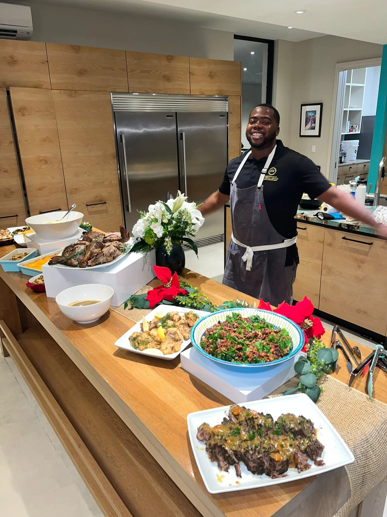 A man smiling in a black shirt and gray apron standing behind a Christmas-themed table filled with food, including roasted meat and various side dishes, in a modern kitchen.