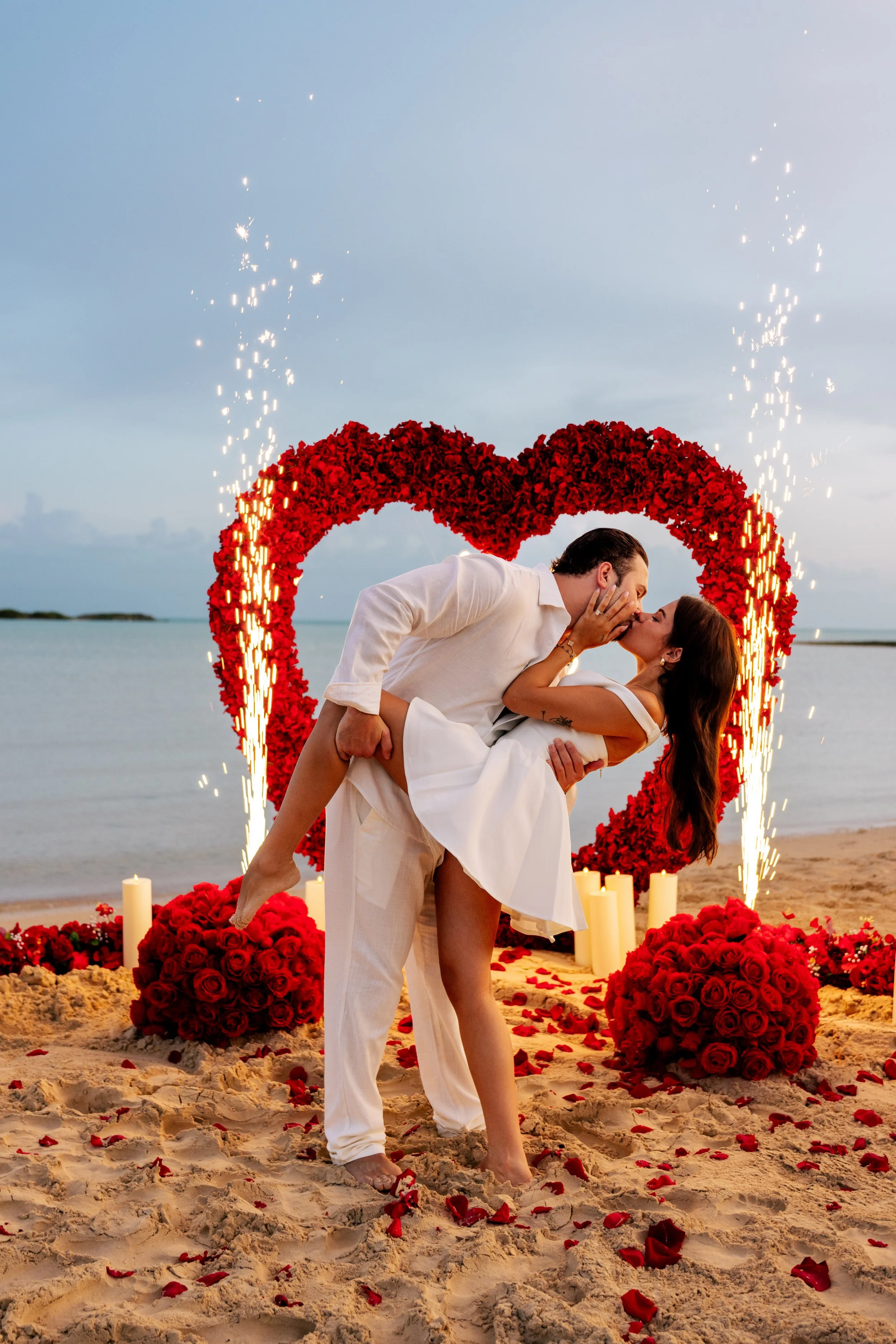 Couple in wedding attire kissing on the beach with a large red heart made of roses behind them, surrounded by candles and scattered rose petals on the sand at sunset.