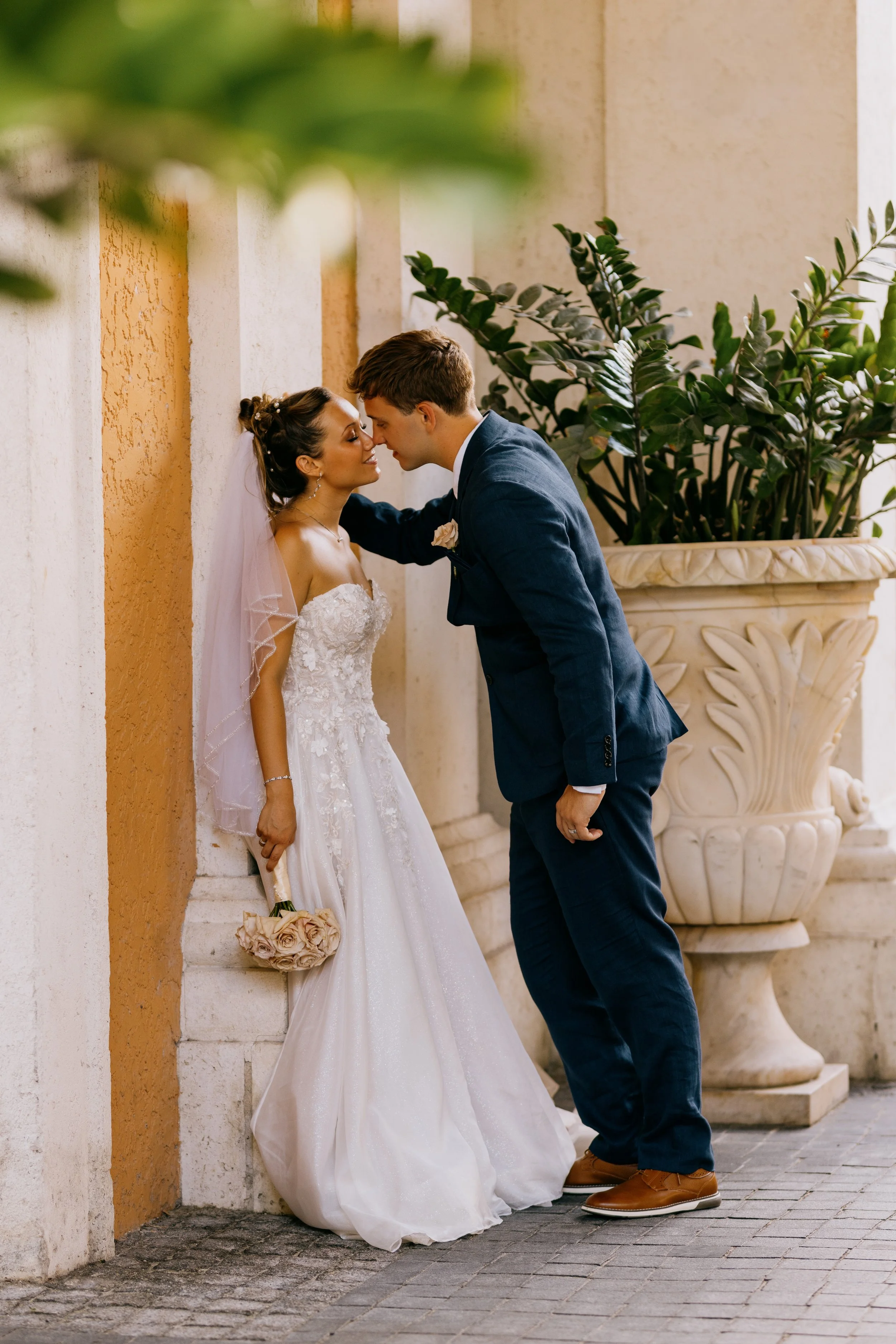 A bride and groom touching foreheads in front of a textured wall with a large potted plant nearby, during their wedding.
