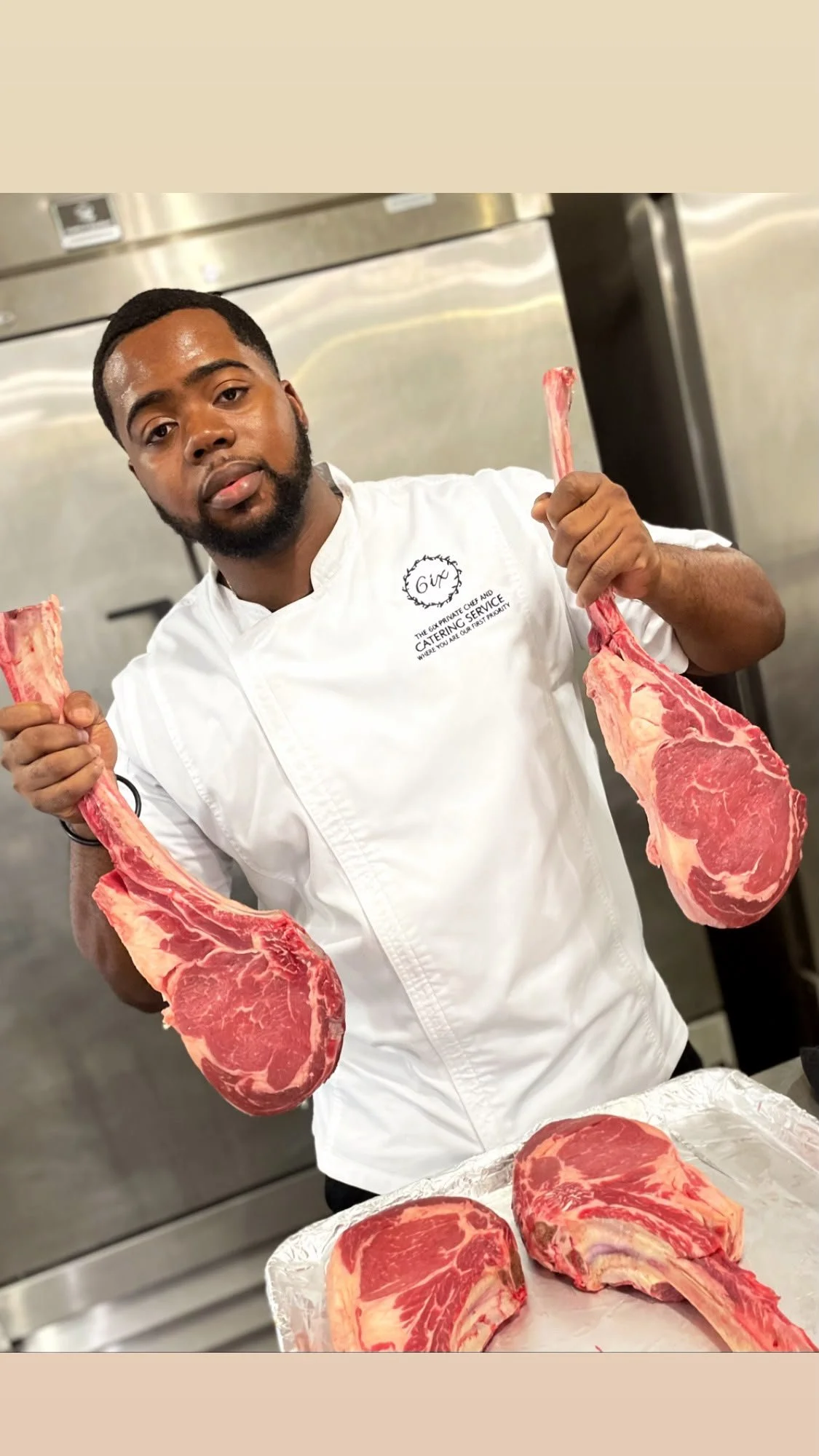 A man in a chef's uniform holding two large raw steaks. There are more steaks on a tray in front of him.