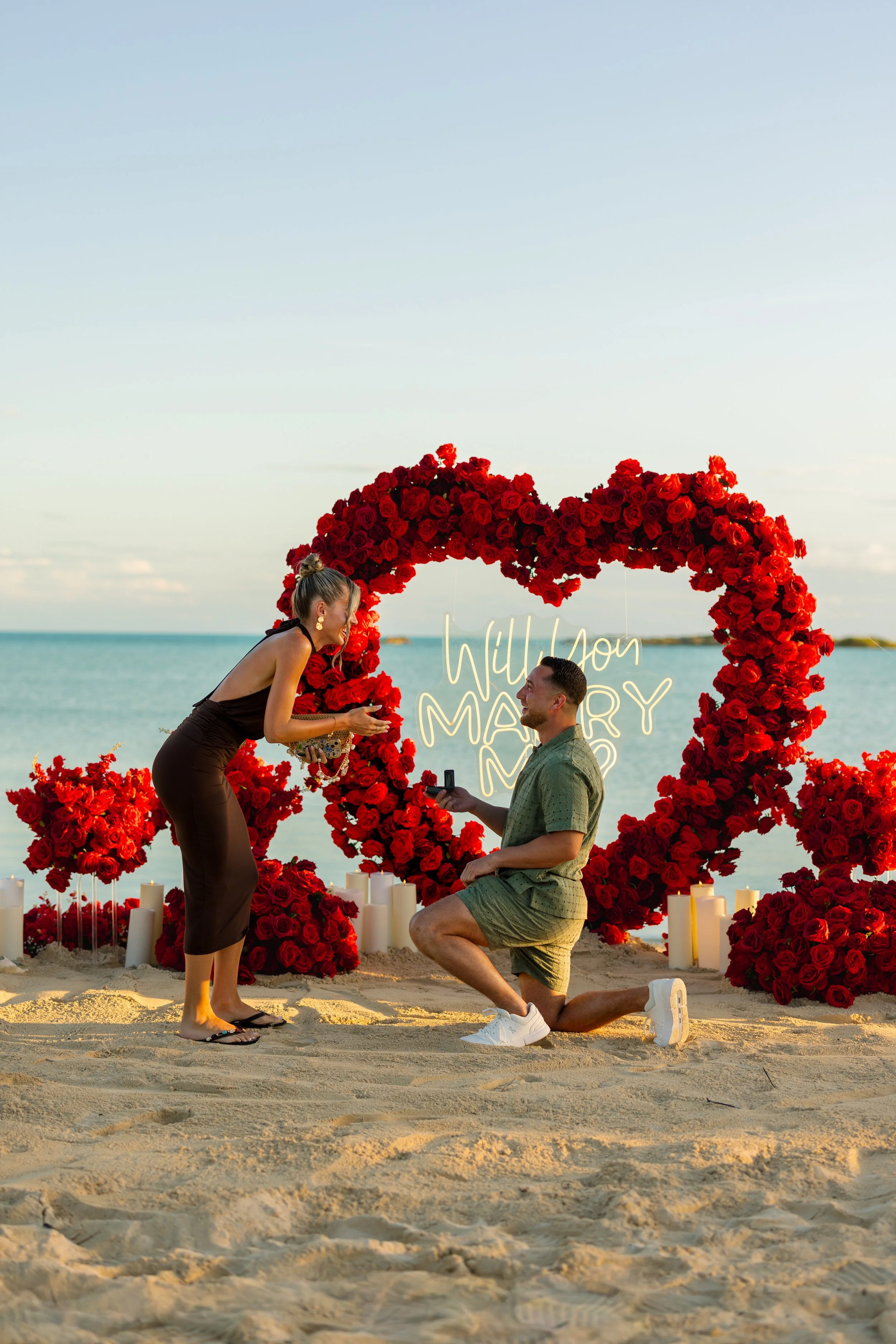 A man proposes marriage to a woman on a beach during sunset. They are surrounded by a floral heart decoration made of red roses and candles, with the word 'Will You Marry Me' lit up in the background.