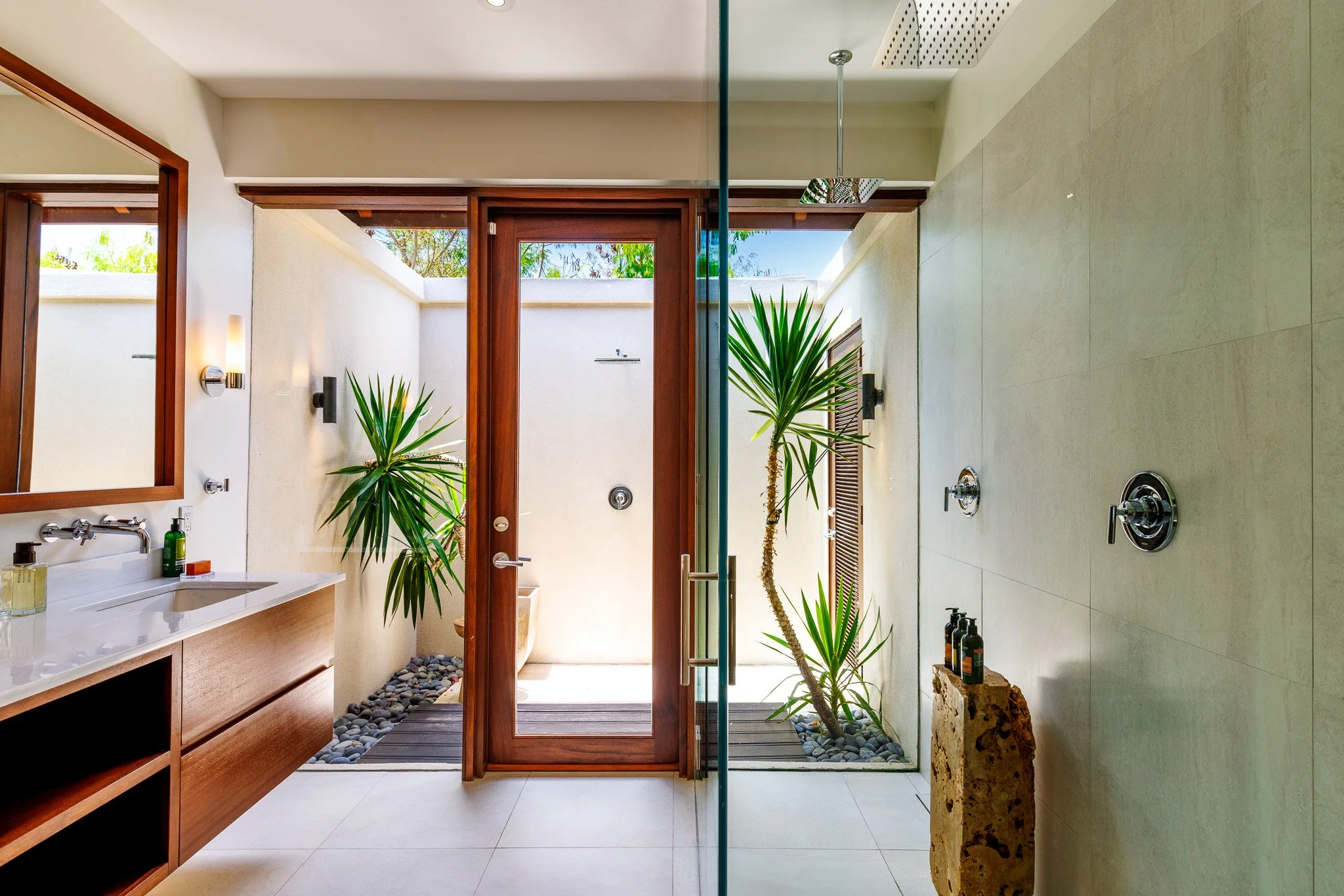 Modern bathroom with a glass shower, wooden accents, potted plants, and outdoor view through glass door.