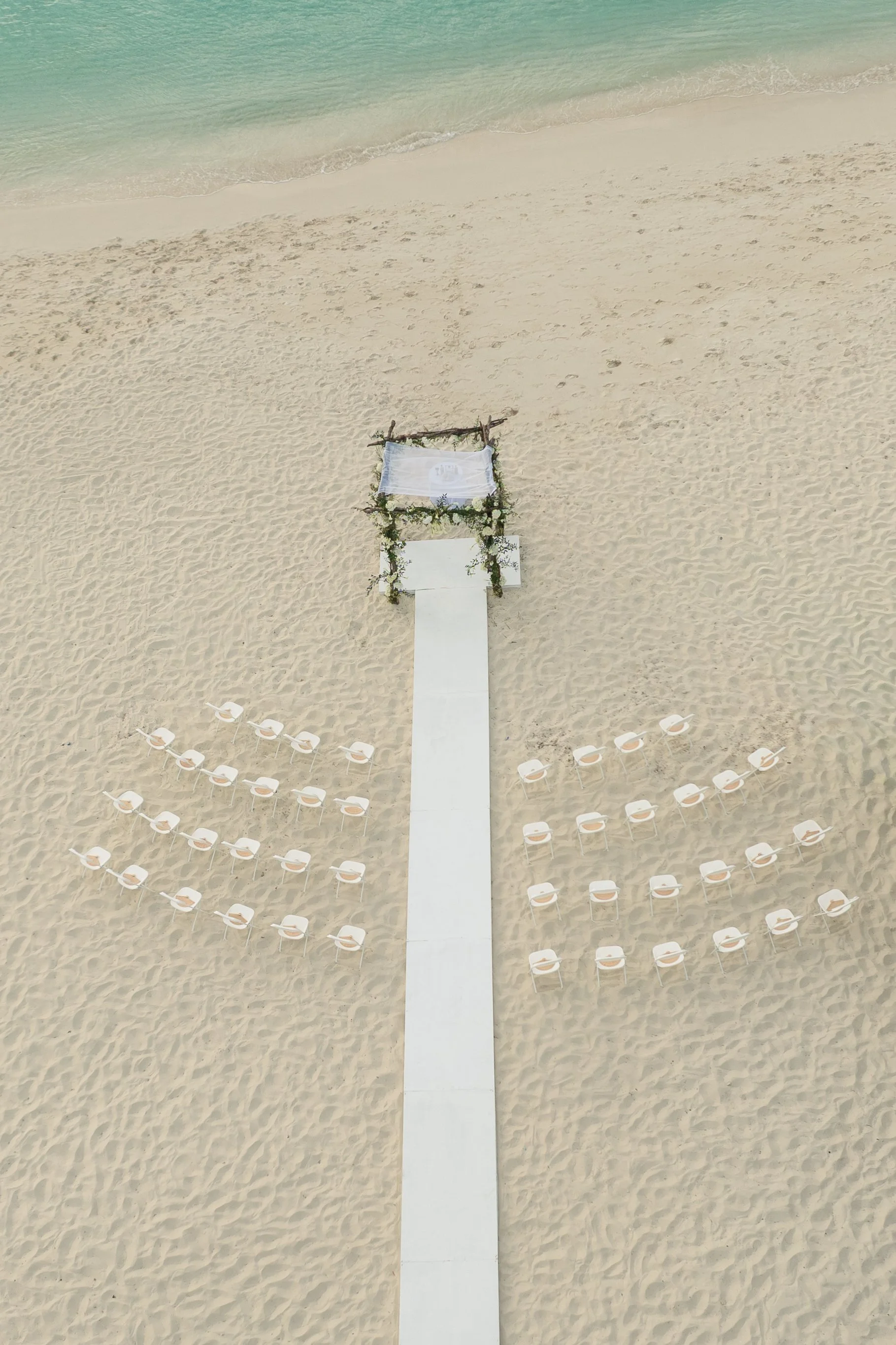 A white aisle runner leading to a wedding arch decorated with greenery on a sandy beach, with rows of white chairs on either side facing the aisle.