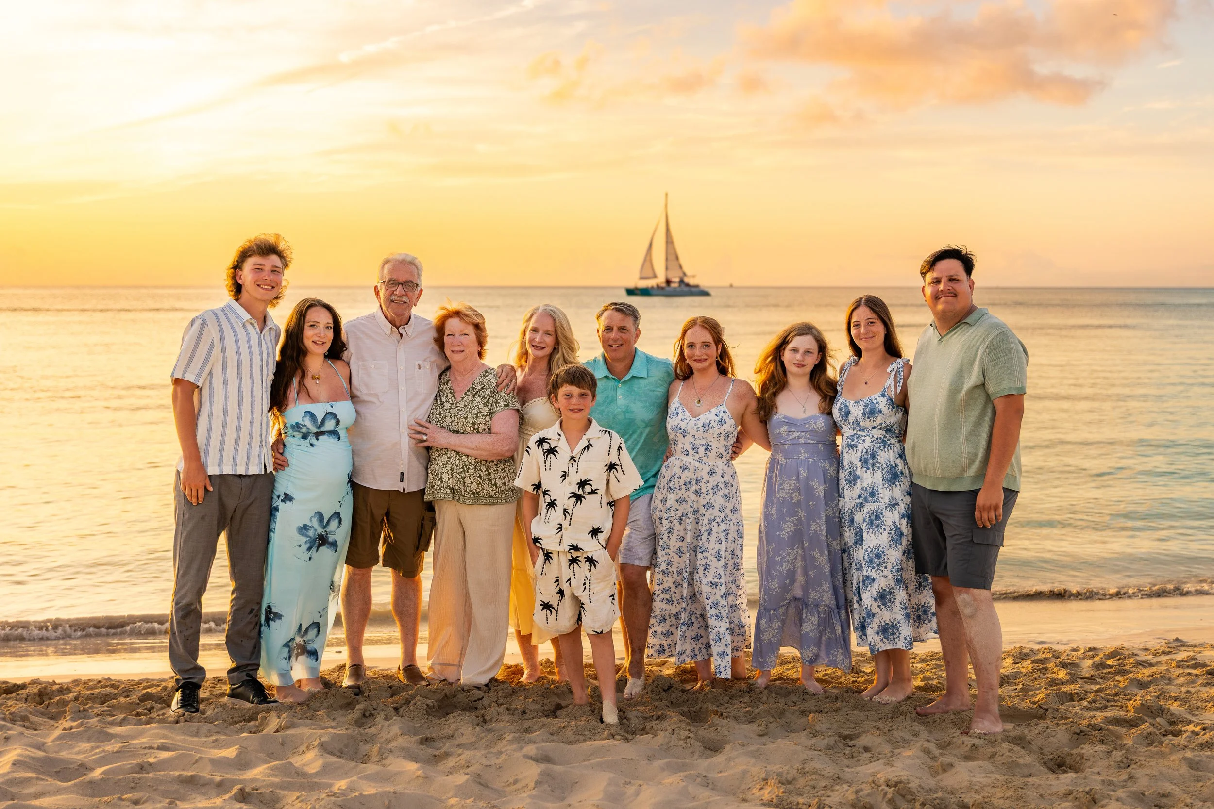 A multigenerational family, including children, adults, and elderly, standing on a beach at sunset, smiling at the camera with a sailboat in the background.