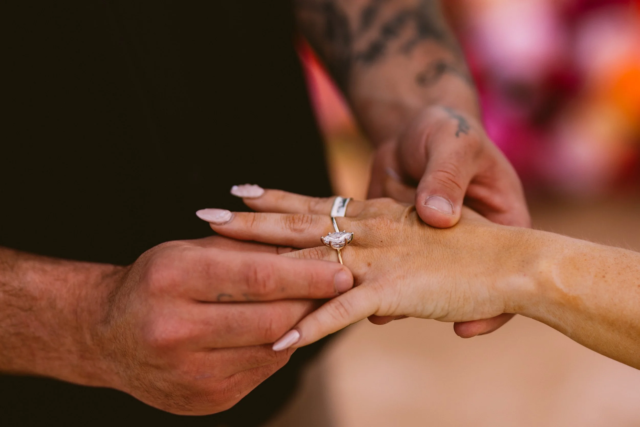 A close-up of three hands, two with visible rings, gripping each other, with tattoos on the upper arm and a colorful background.