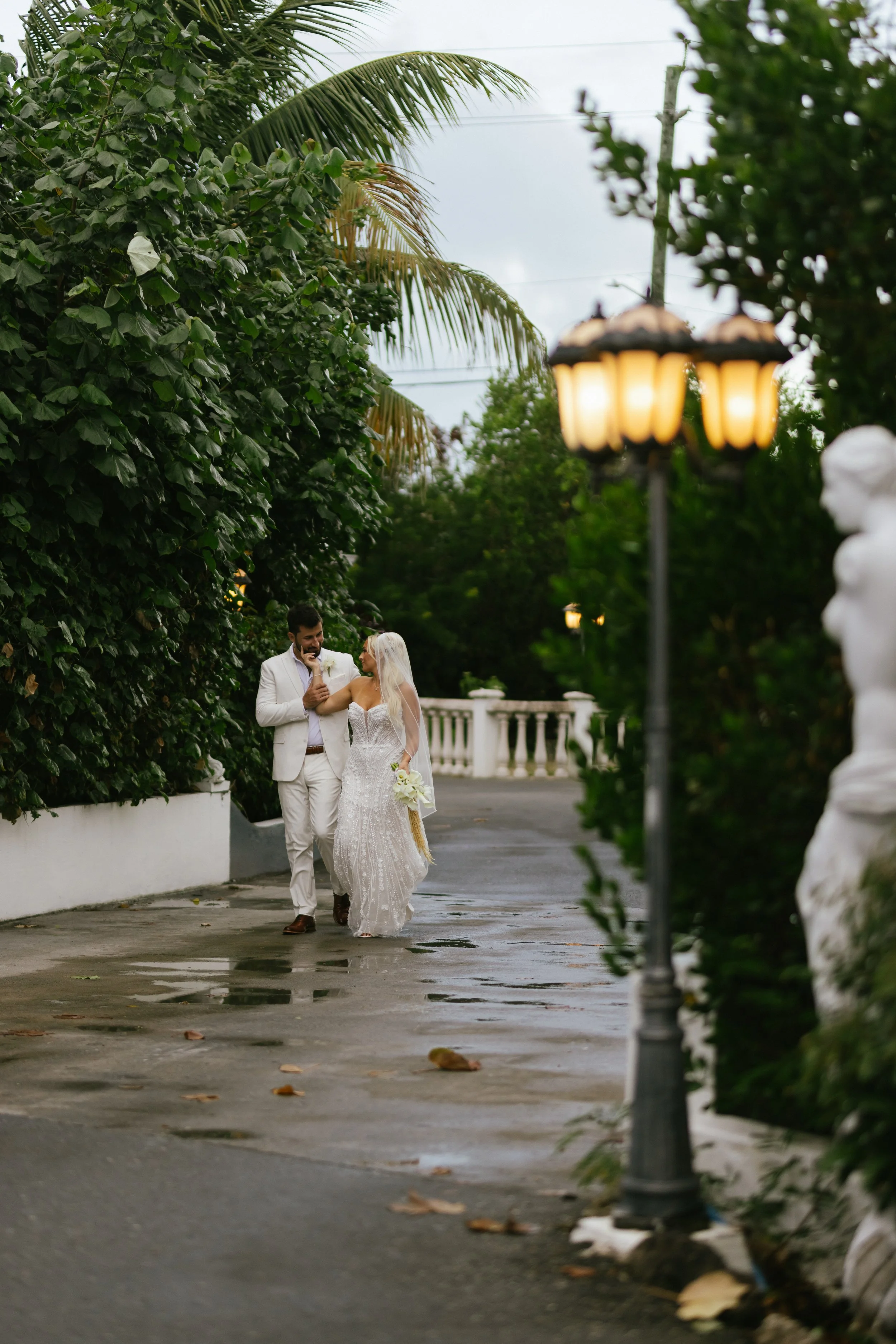 A bride and groom walking outdoors in the rain, surrounded by green foliage and street lamps.