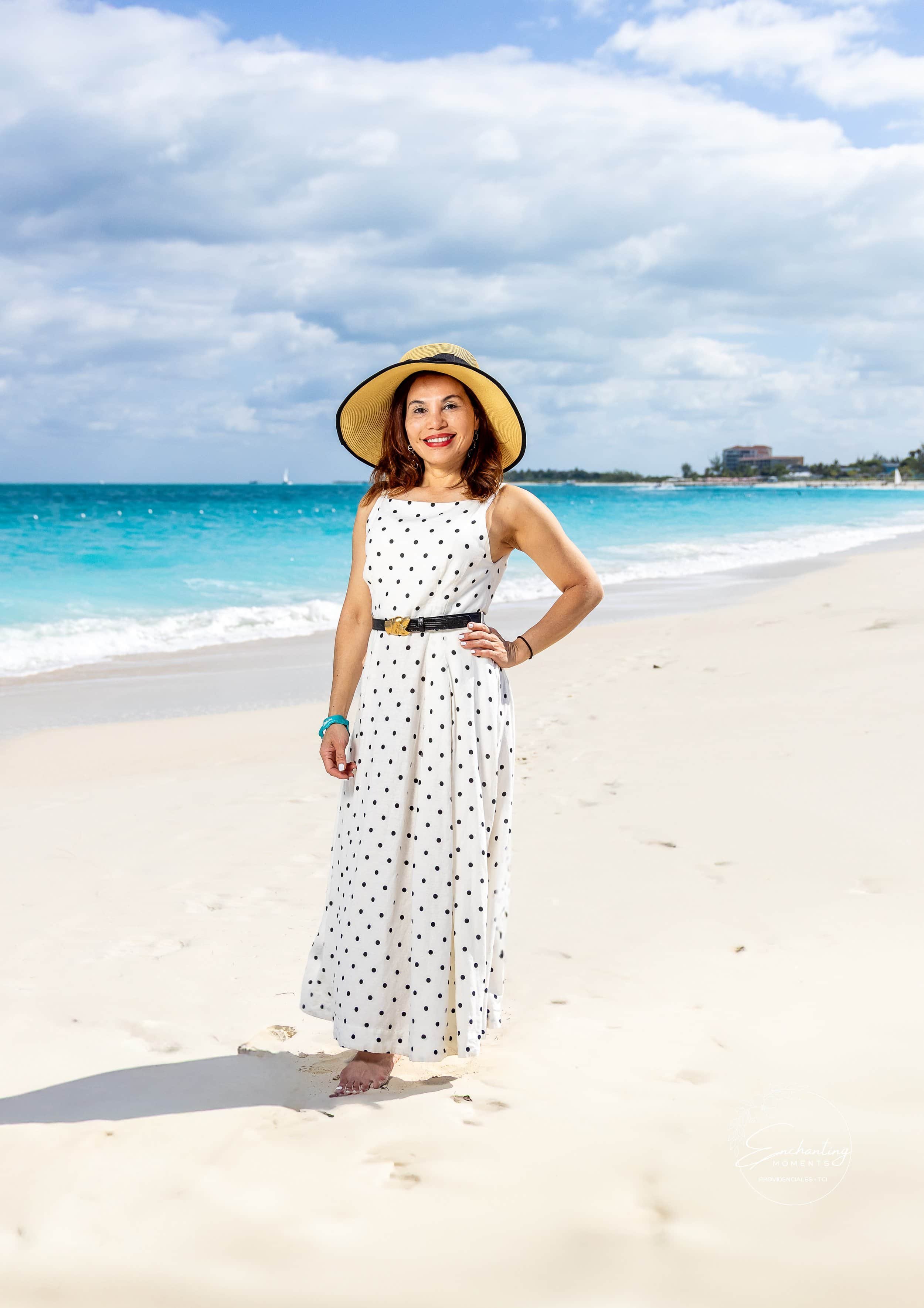 Woman in a polka dot dress and wide-brim hat smiling on a sandy beach with ocean and cloudy sky in the background.