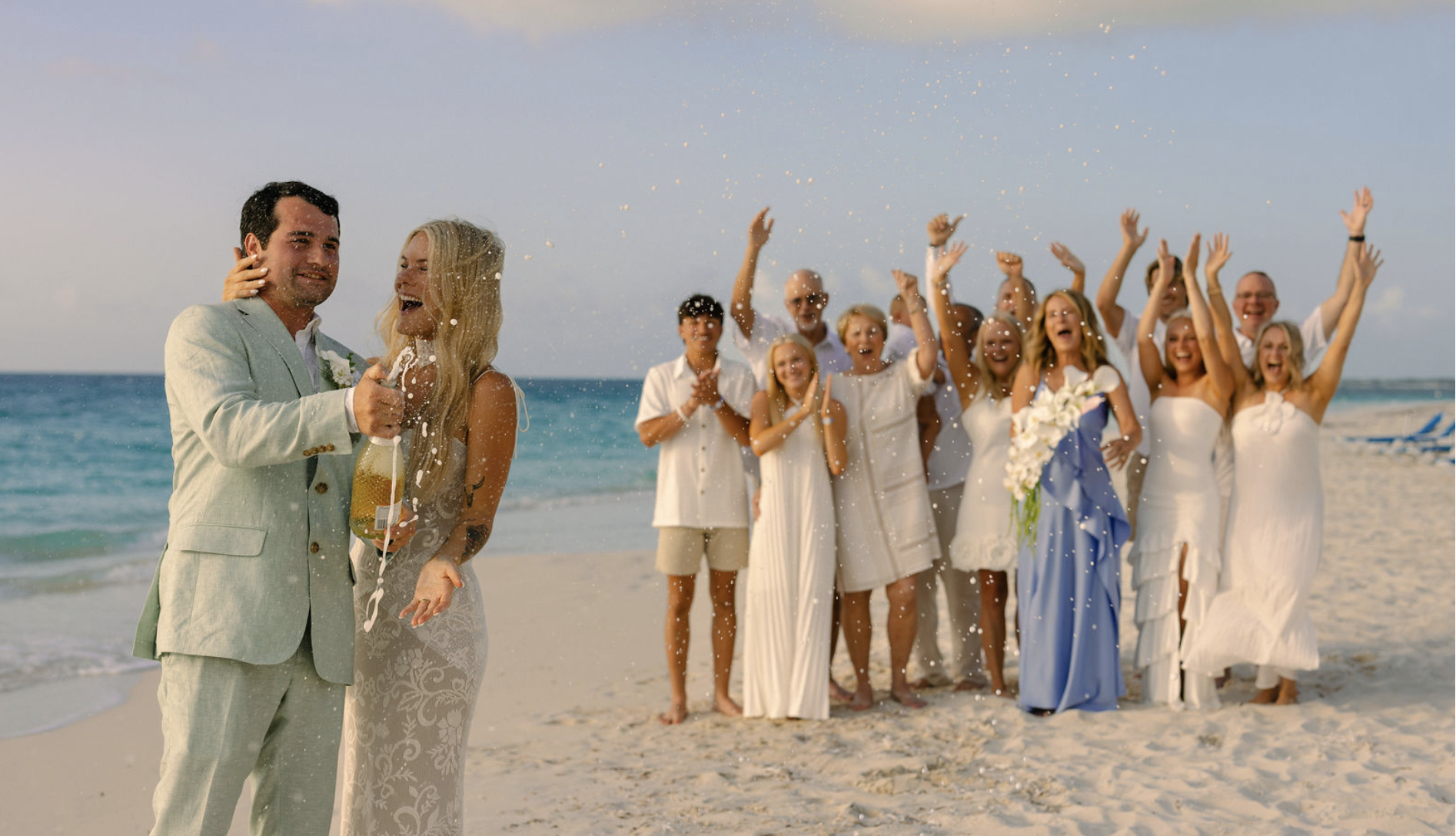 A wedding celebration on the beach with a bride and groom in the foreground, the groom opening a bottle of champagne, and a group of people in white dresses and shirts cheering and celebrating in the background.