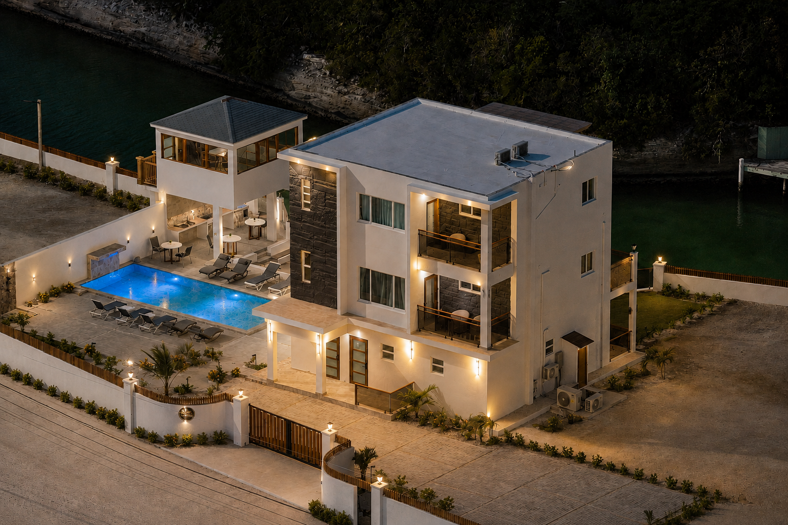 A modern three-story house with a swimming pool, lounge chairs, outdoor dining areas, and balconies, surrounded by a white fence with decorative lighting, near a body of water at night.