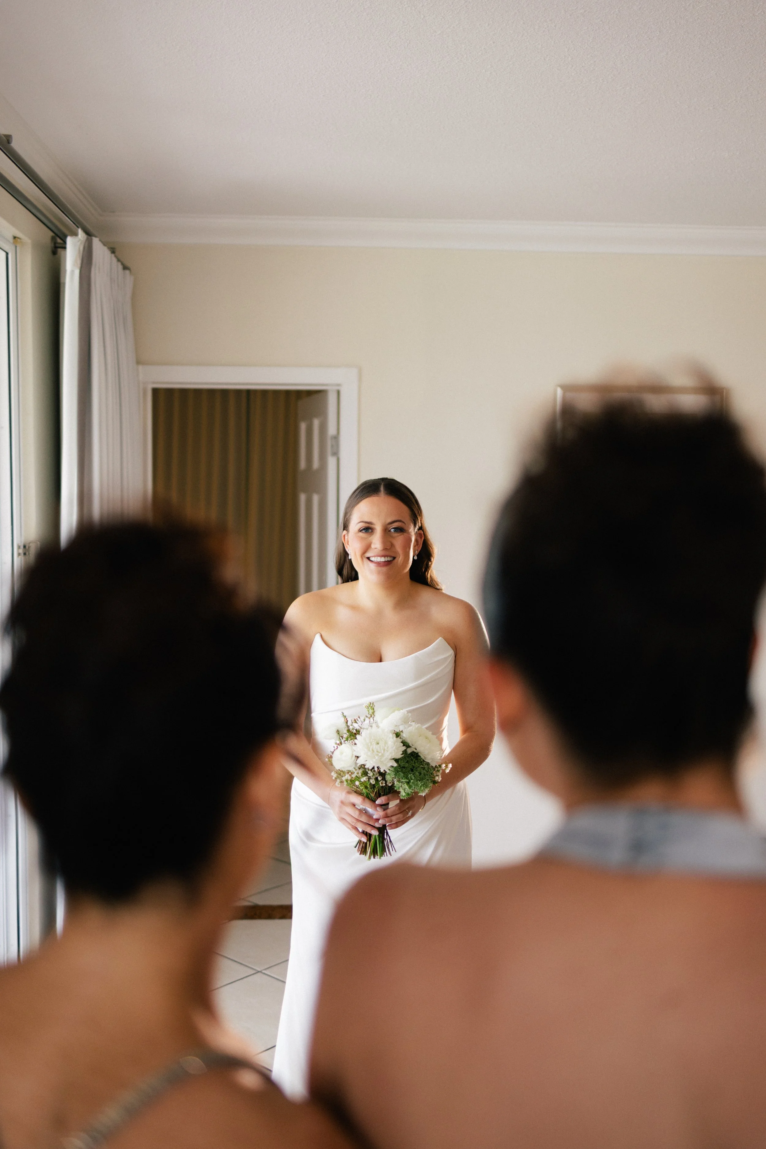 A smiling bride in a white wedding gown holding a bouquet of white flowers, looking towards the guests in a room with cream walls and white curtains.
