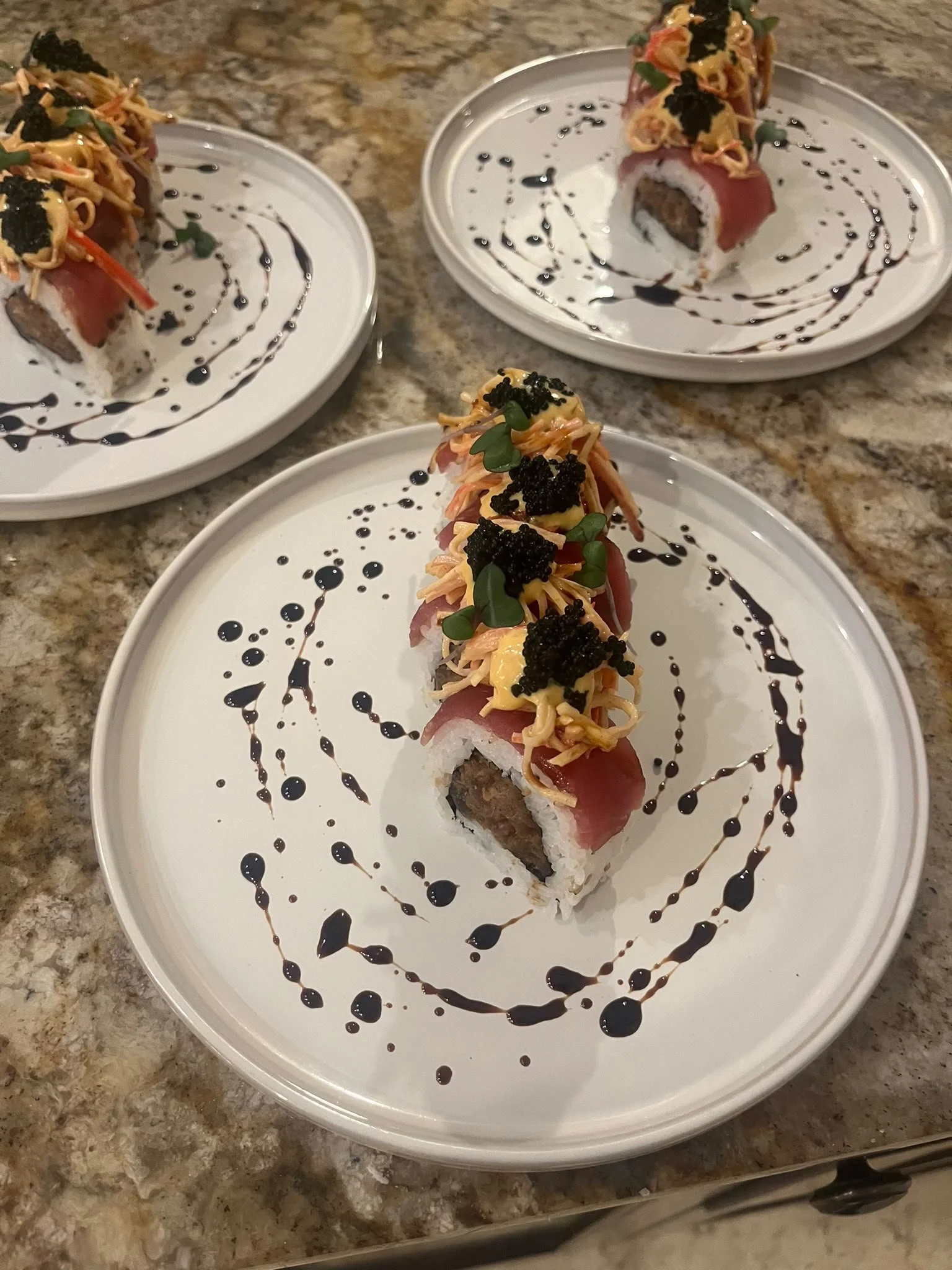 Close-up view of sushi rolls topped with shredded cheese, black fish roe, microgreens, and drizzled with dark sauce, served on white plates on a granite countertop.