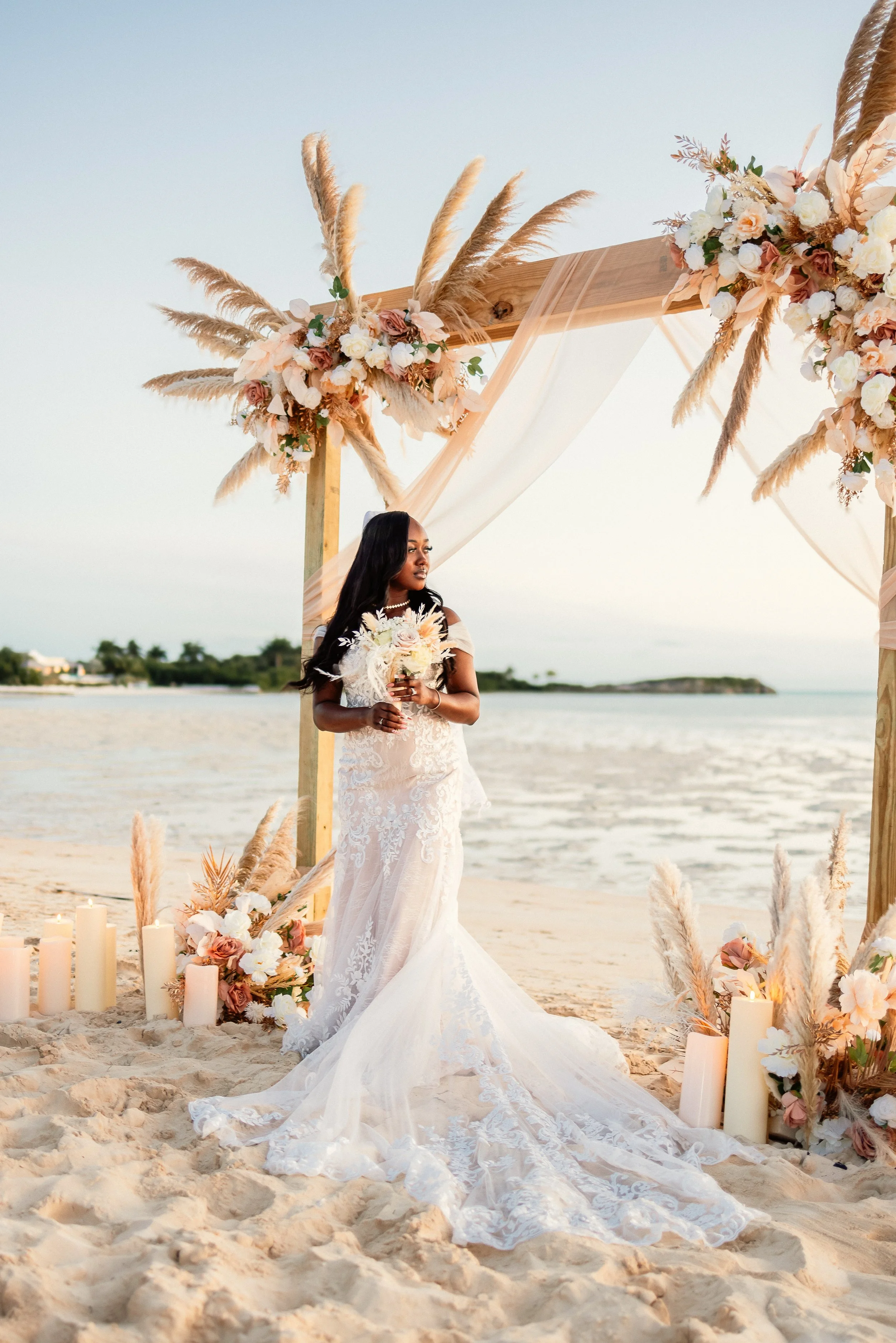 Bride in a wedding dress holding a bouquet, standing on a sandy beach under a decorated arch with flowers and candles, during sunset.