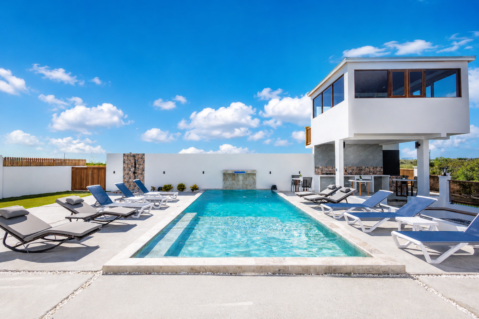 Modern backyard with a rectangular swimming pool, lounge chairs, a water feature, and a two-story white house with large windows under a blue sky with scattered clouds.