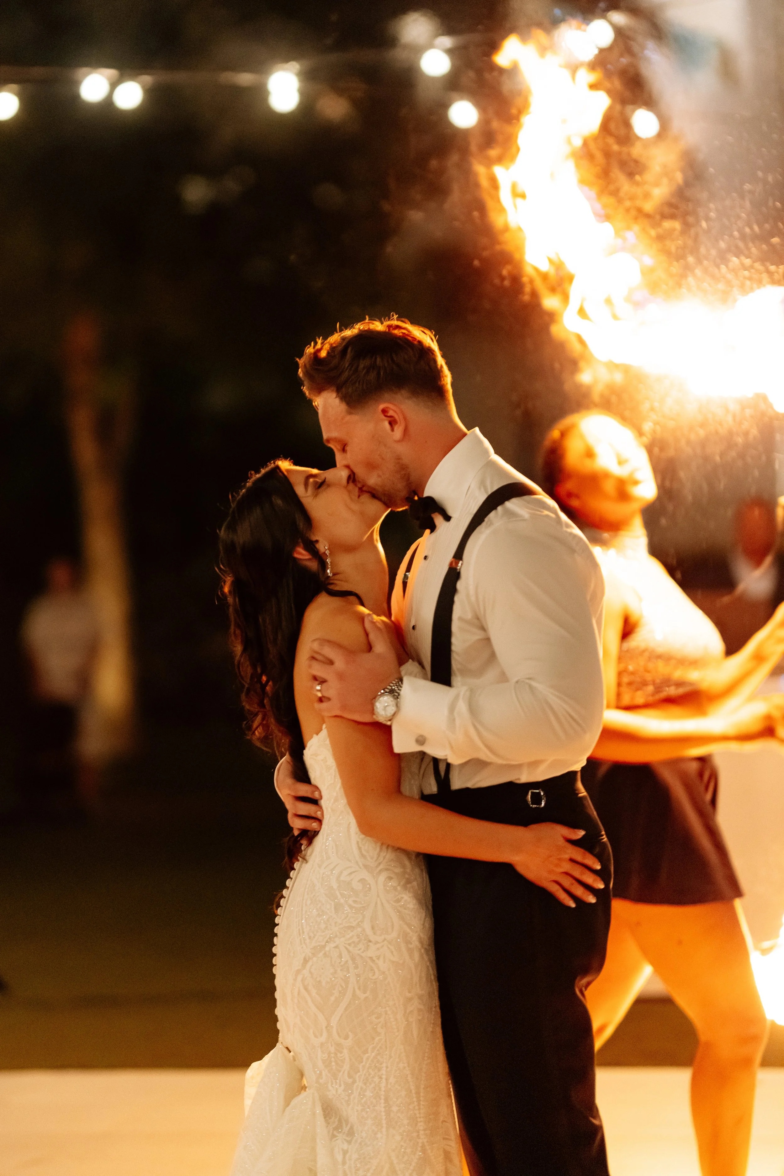 A bride and groom are sharing a kiss while dancing at their wedding reception, with fire dancing in the background.