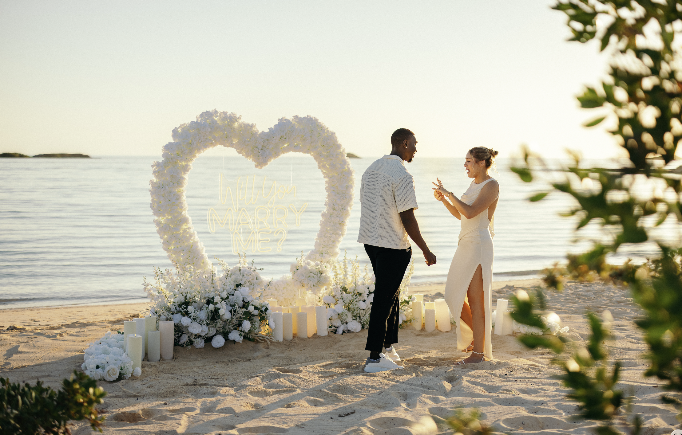 A man and woman get engaged during a beach wedding ceremony with a floral heart and candles, at sunset, with the ocean in the background.