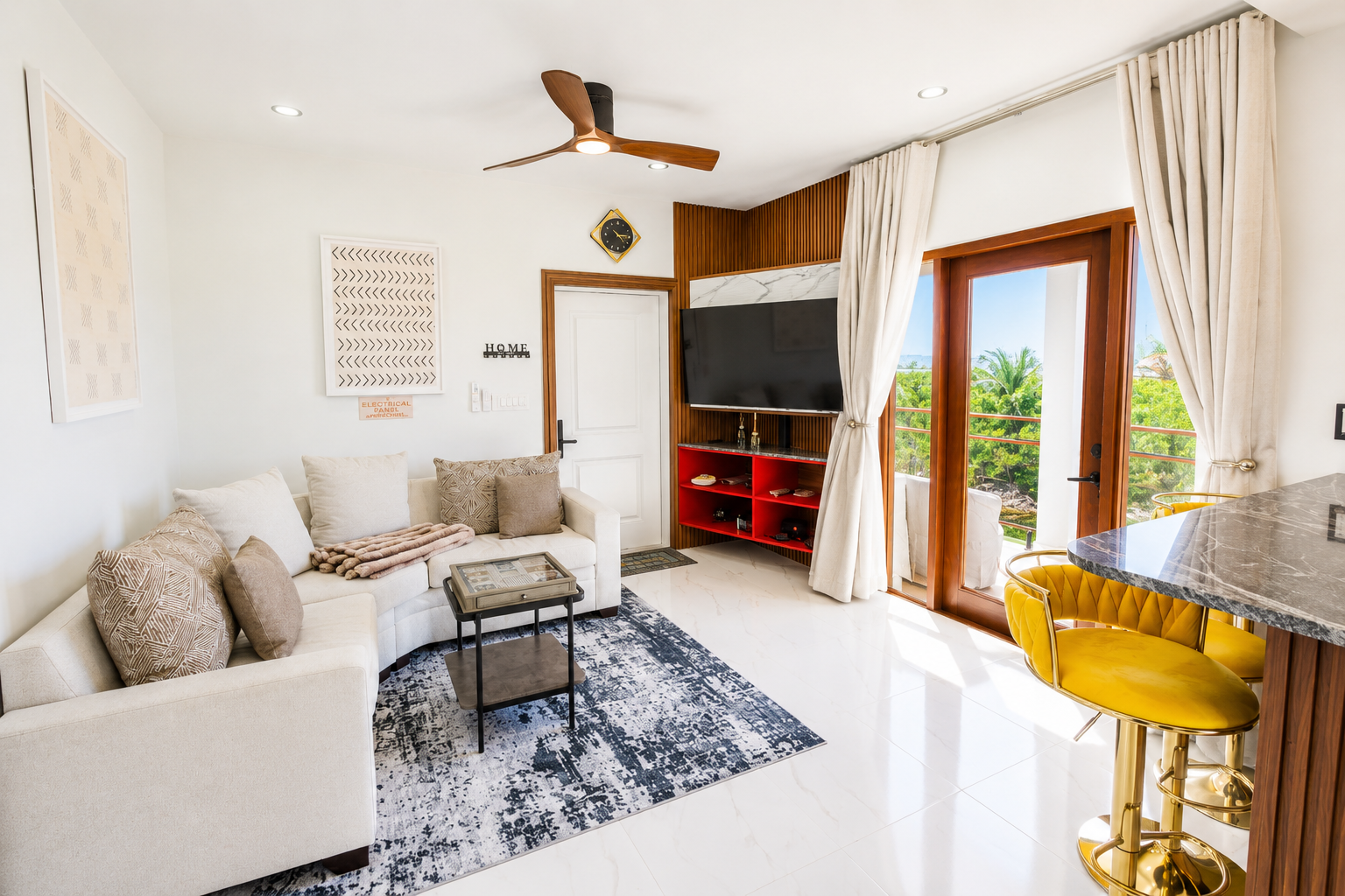 Living room with white sofa, patterned pillows, a small black coffee table, a navy and white rug, wall art, a TV on a wooden accent wall, a balcony with glass doors, cream curtains, and a yellow bar stool.