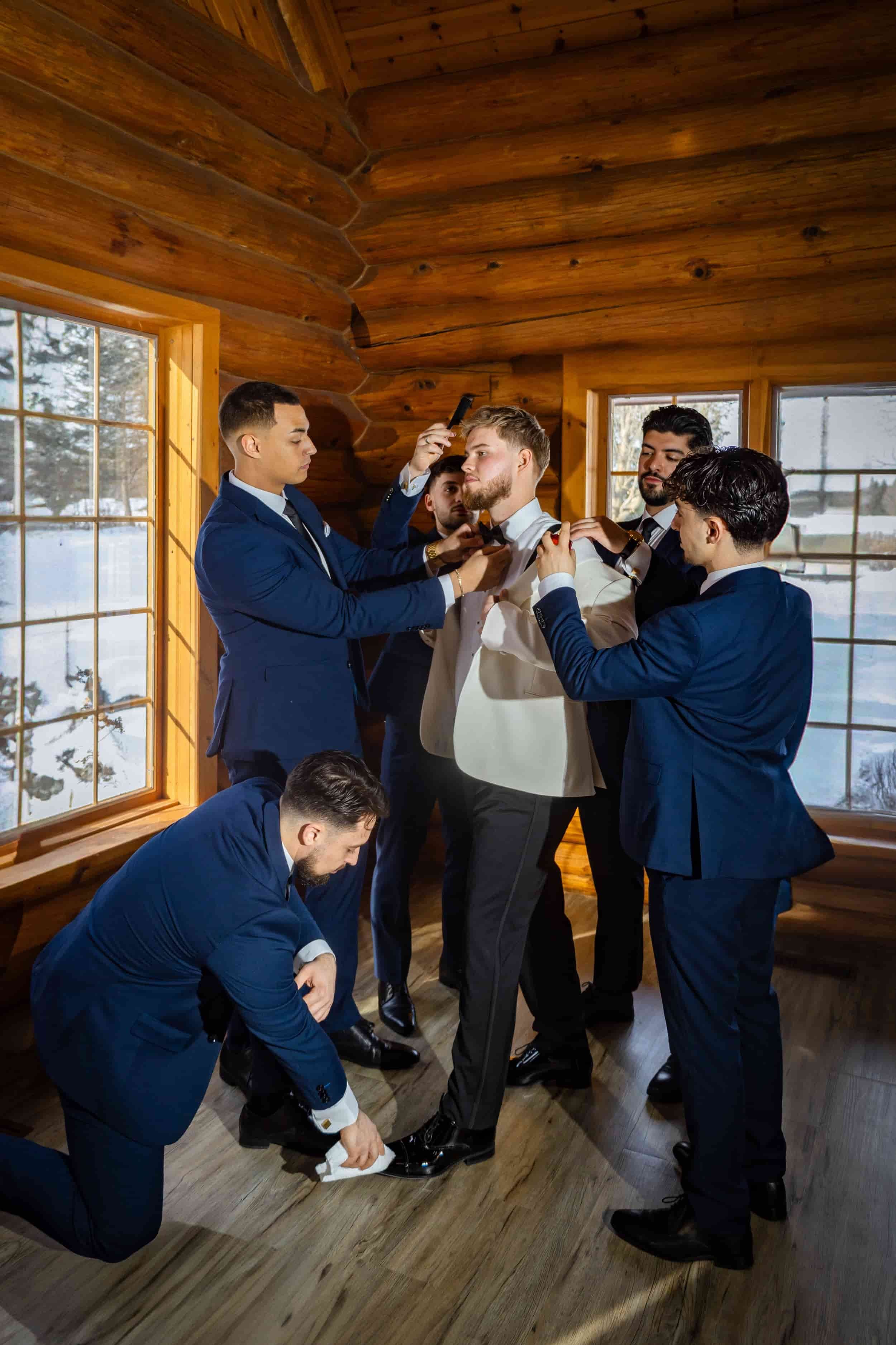 Group of men in suits helping groom prepare for his wedding in a wooden cabin.