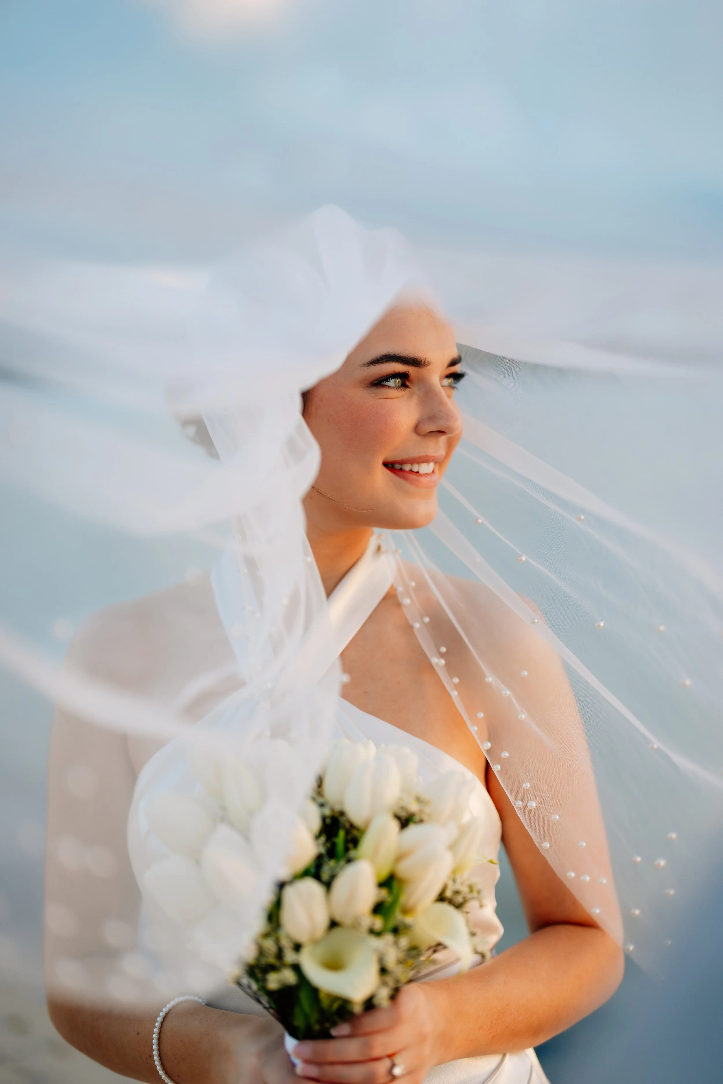 A smiling bride holding a bouquet of white flowers, wearing a wedding dress and veil with a light blue sky in the background.