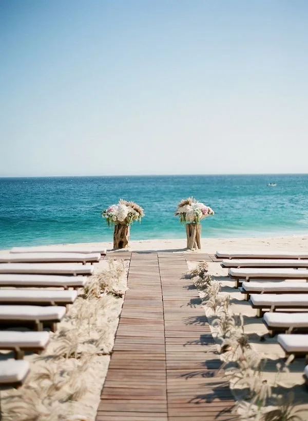 Beach ceremony setup with an aisle, guest seating and a floral arch beside turquoise water
