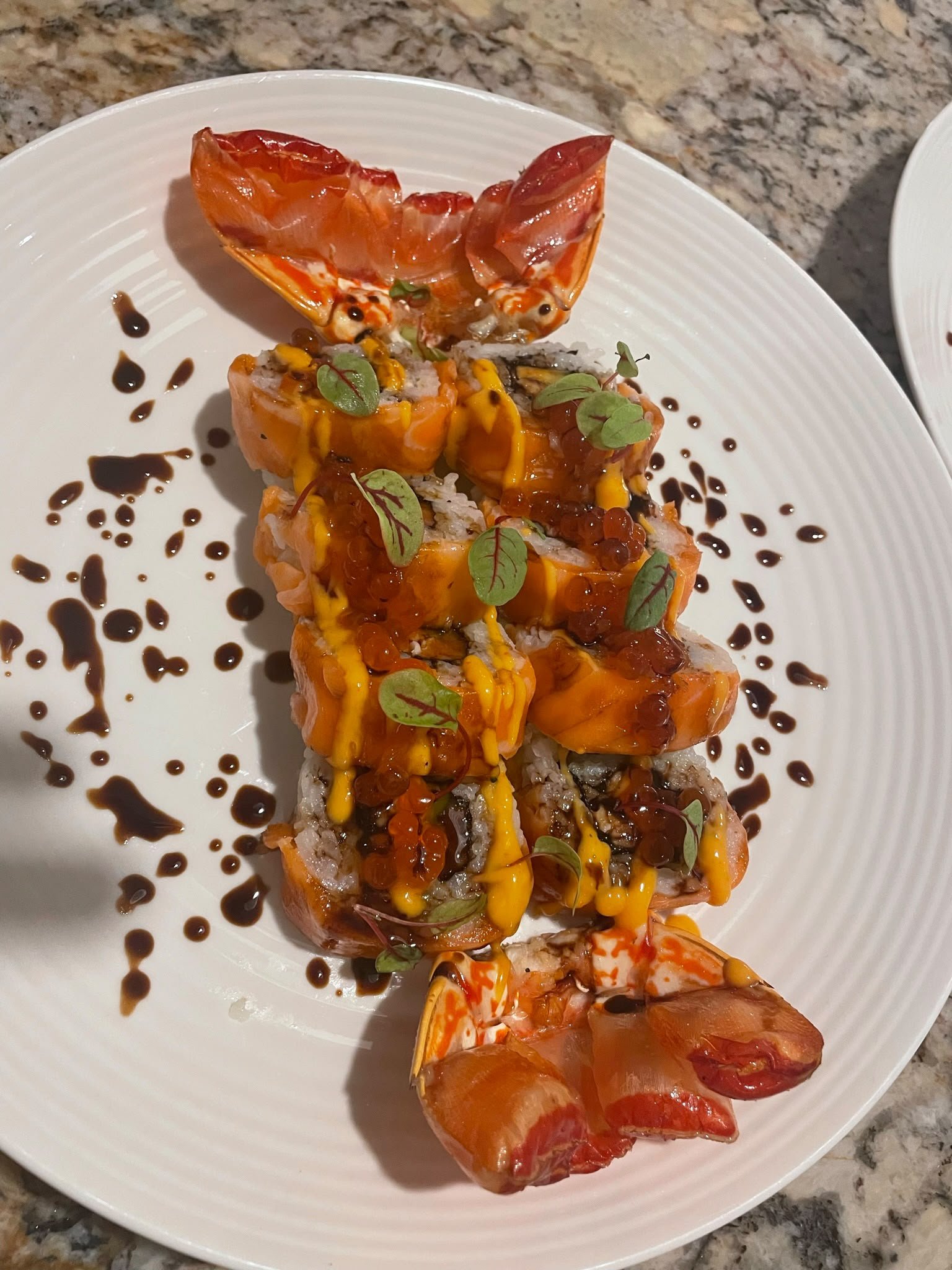 Sushi roll topped with sauces, microgreens, and garnished with sauces on a white plate set on a granite countertop.