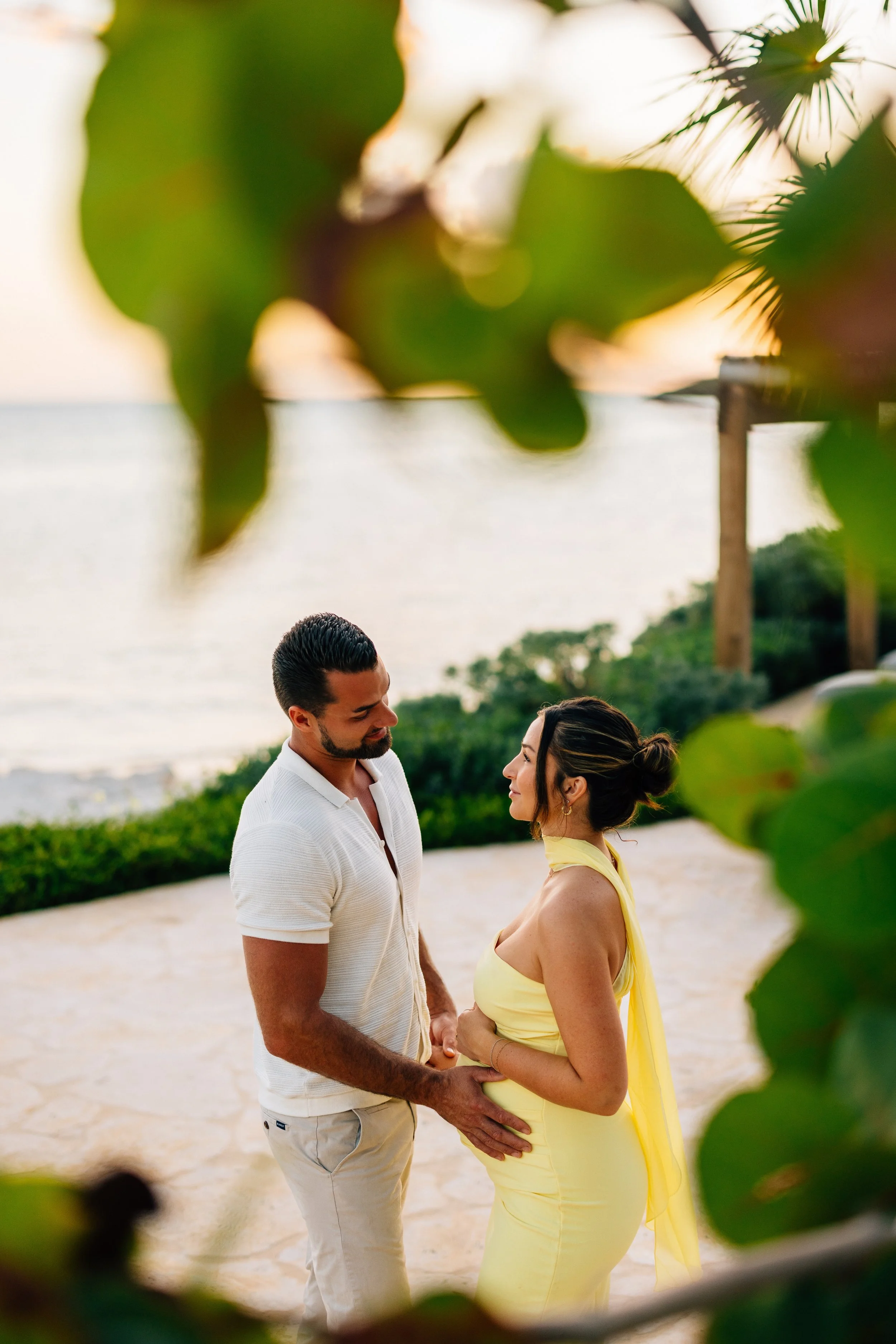 A couple standing close and holding hands on the beach, framed by green leaves, with the sea and sky in the background during sunset.