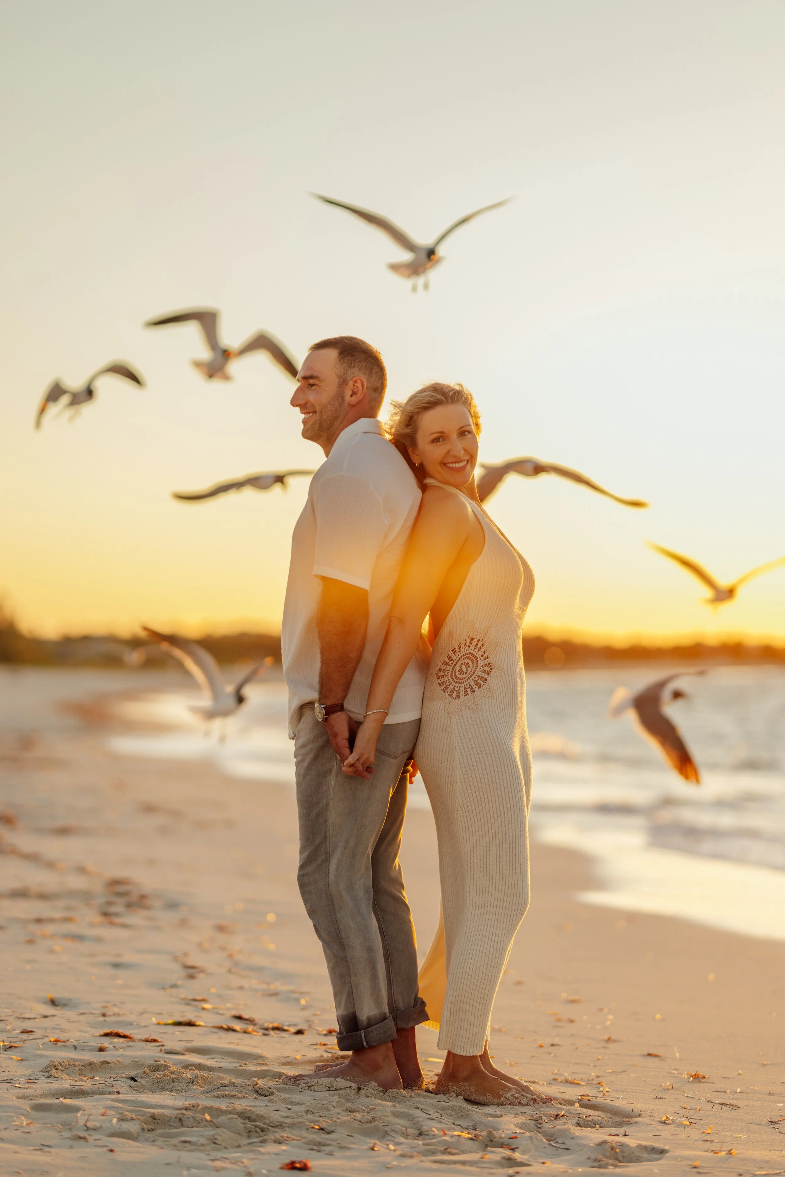 A smiling couple stands back to back on a beach at sunset, holding hands, with seagulls flying around them and the ocean in the background.