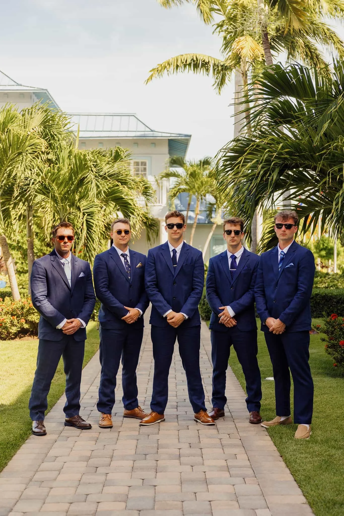 Five men dressed in blue suits and sunglasses standing on a brick pathway lined with palm trees and greenery, with a large house or hotel in the background.