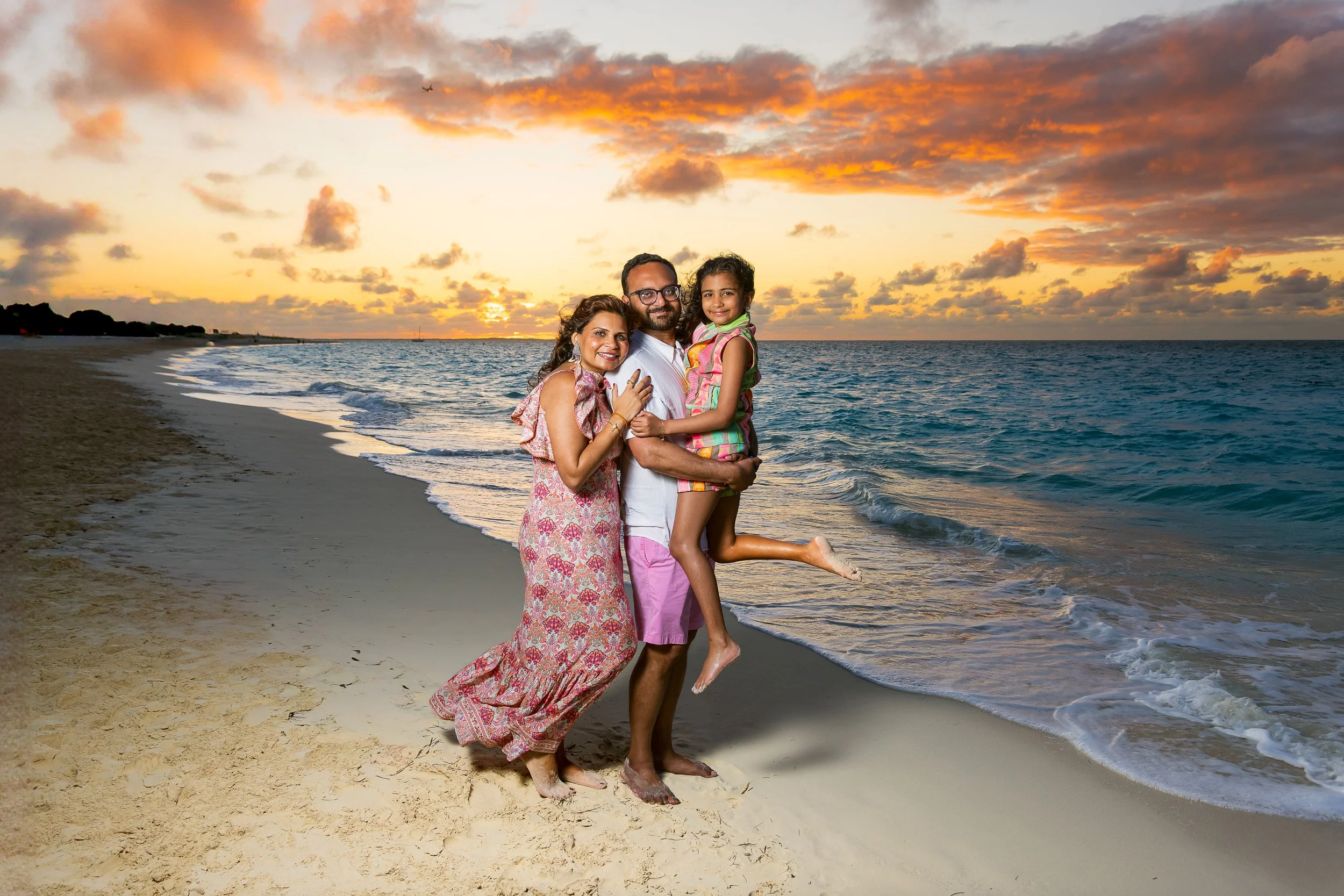 A family of three on a beach at sunset, with a woman in a floral dress, a man in pink shorts, and a young girl in a colorful dress, all smiling and holding each other.