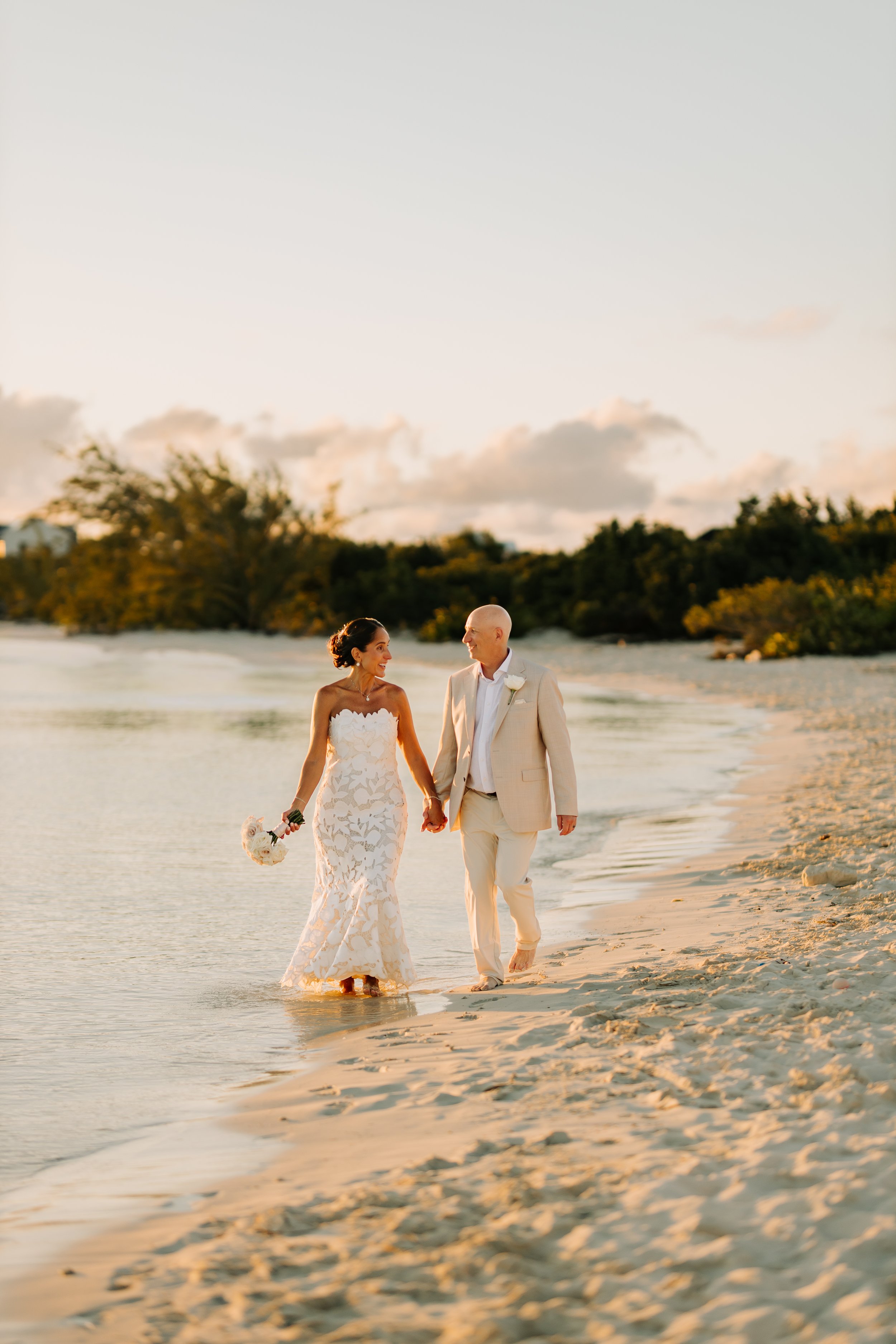 A bride and groom walking hand in hand along the beach at sunset, smiling at each other, with the bride holding a bouquet of flowers.