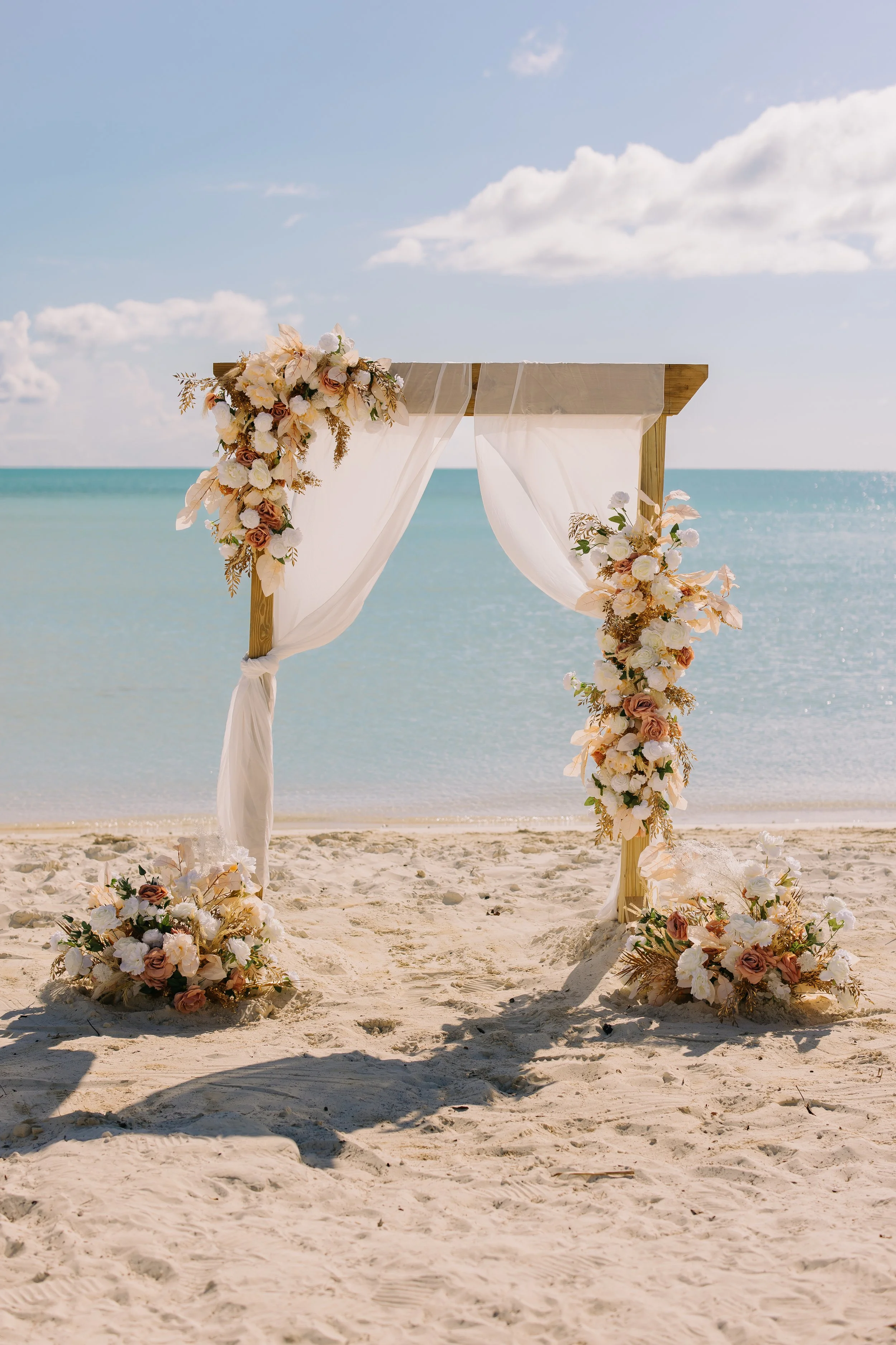 Beach wedding arch decorated with flowers, draped with white fabric, set on sandy beach by the ocean.