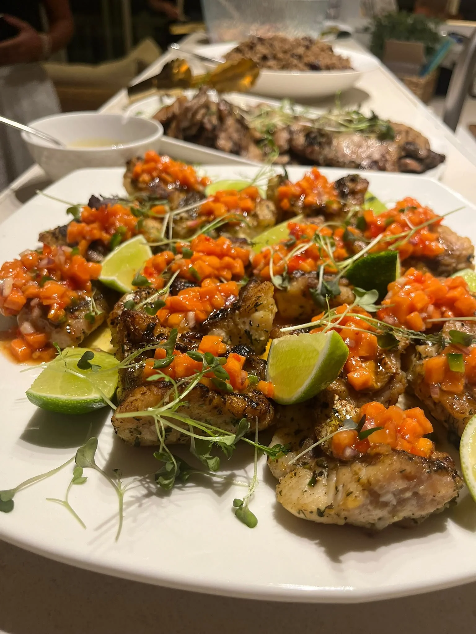 Close-up of a white plate with grilled chicken topped with a red and orange vegetable salsa and garnished with lime wedges and microgreens, with a background of other dishes on a dining table.